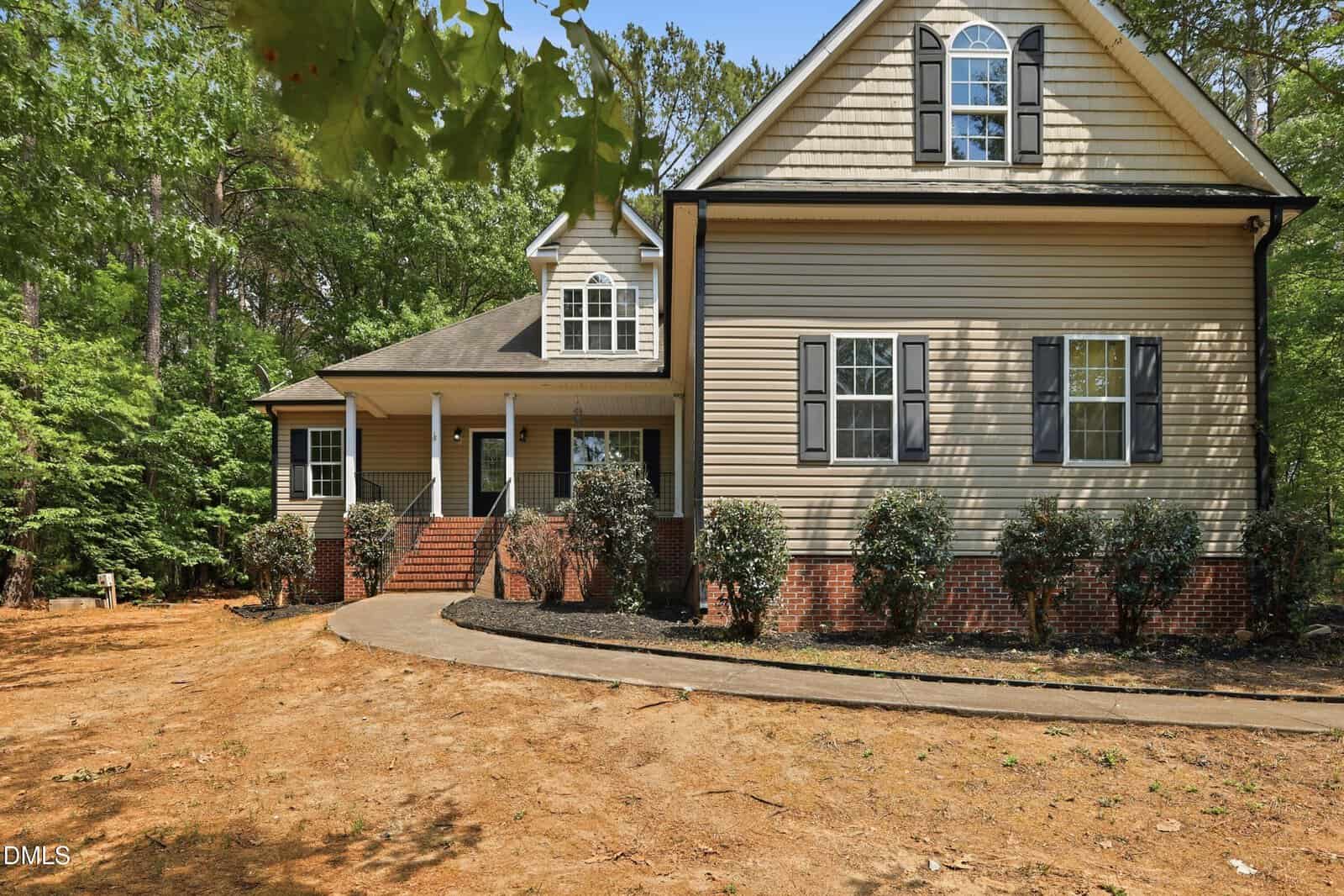 A beige two-story house with black shutters and a brick foundation sits at 2130 Turquoise Lane in Franklinton. Surrounded by shrubbery, trees, and bordered by woods, it features a curved walkway to a covered front porch and patchy grass in the yard.