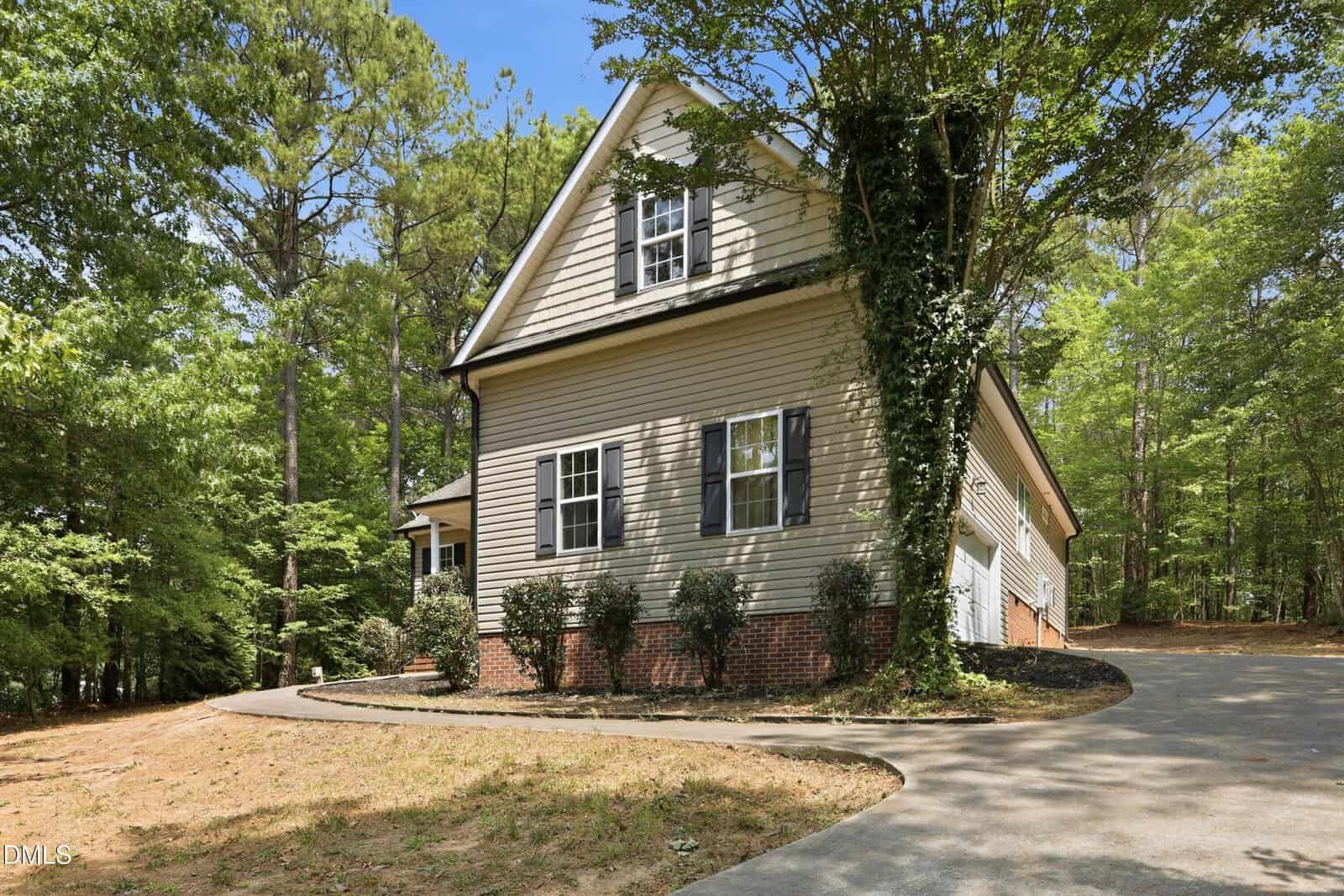 A two-story beige house with white trim, brick foundation, and black shutters sits in a wooded area. A curved driveway leads to a garage on the right side, surrounded by trees and greenery.