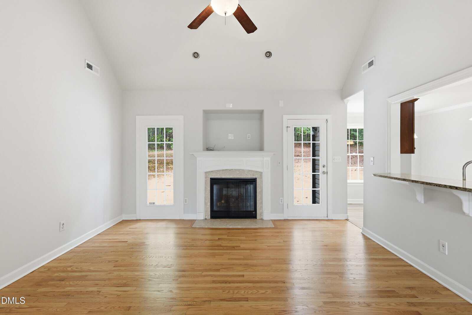 A bright living room with hardwood floors, a white fireplace centered on the back wall, two glass doors on either side, and a ceiling fan above. There are openings to adjacent rooms on the right.