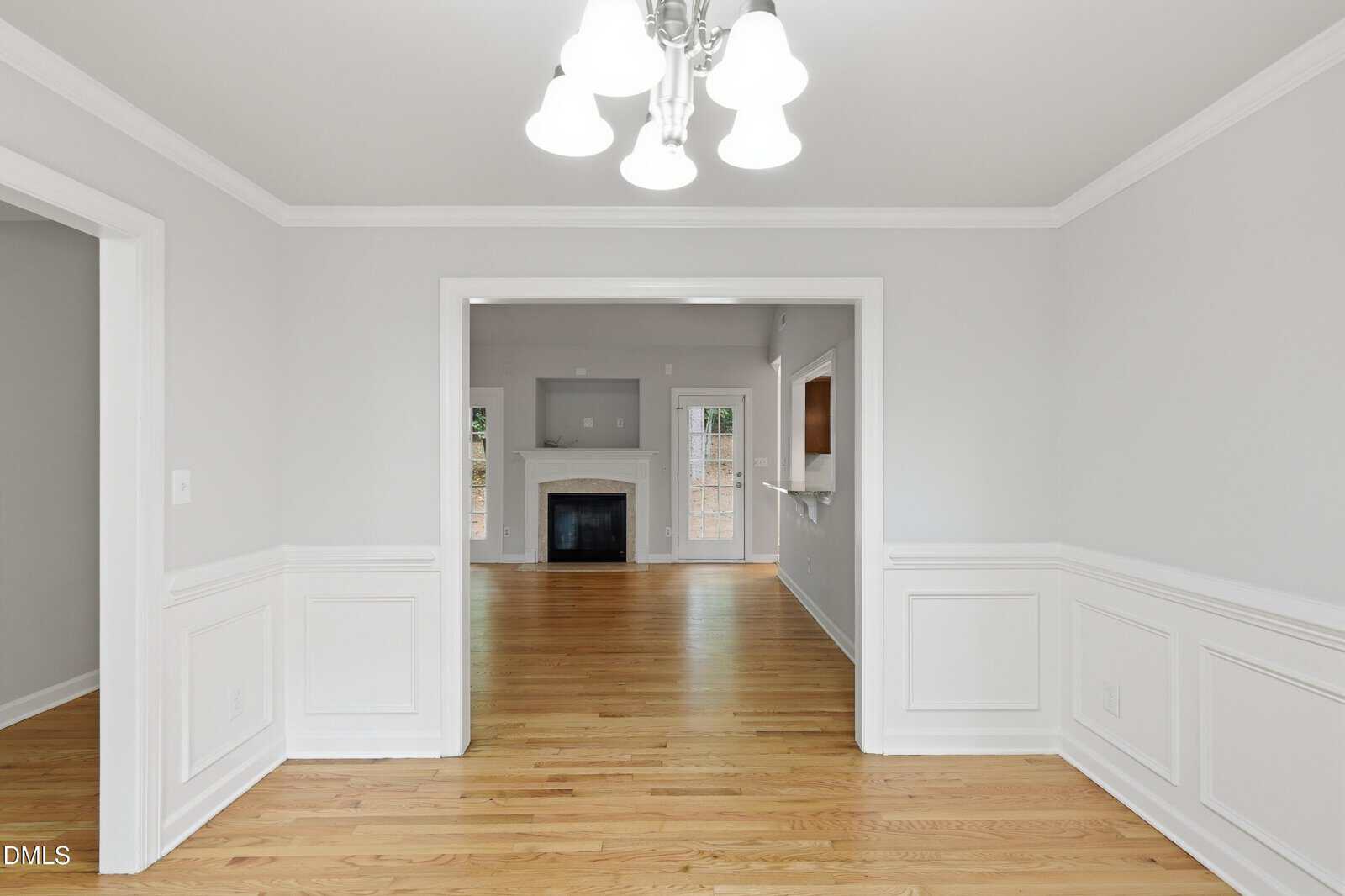 A bright dining room with white wainscoting and light gray walls opens into a living area with hardwood floors, a white fireplace, and glass doors leading outside. Ceiling lights hang above the dining space.