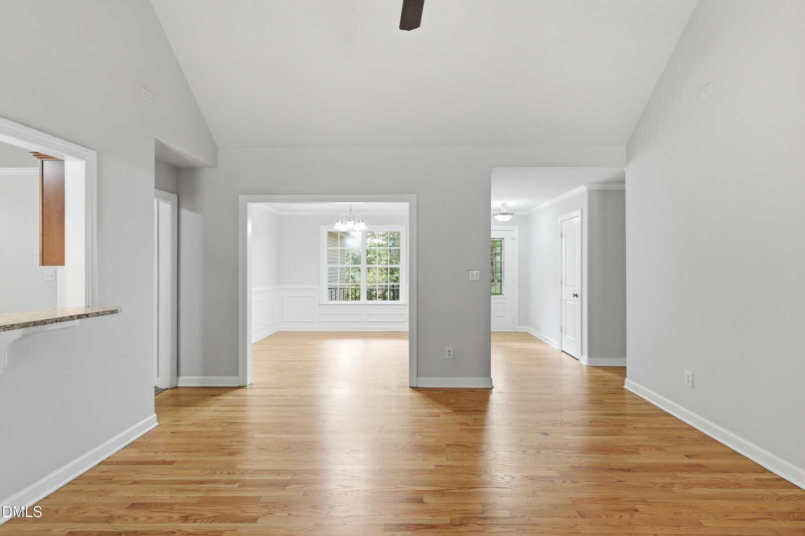 Bright, empty room with light wood floors, white walls, and large windows. There is a pass-through window on the left, a dining area in the center, and a hallway leading to doors on the right.