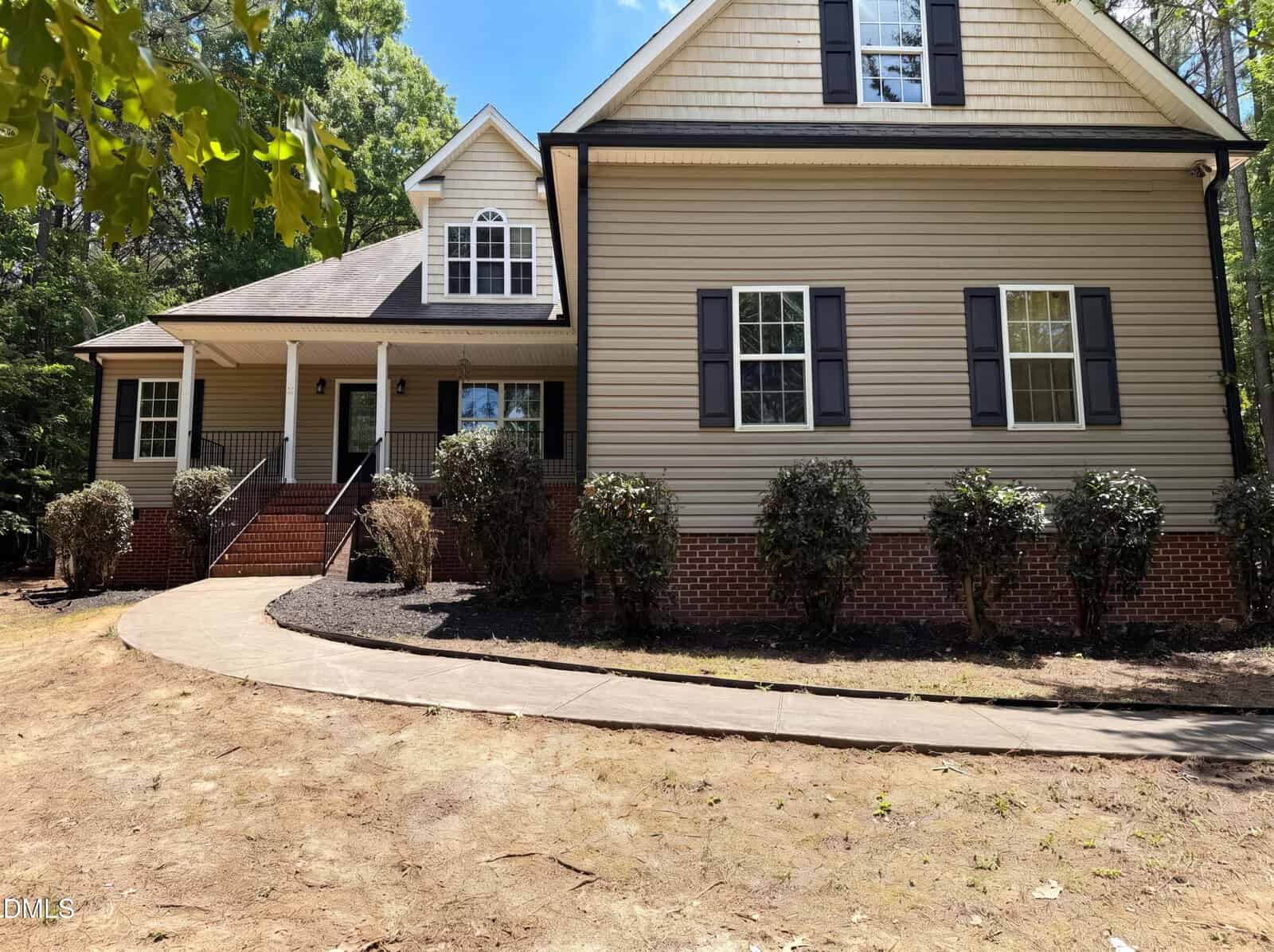 A beige two-story house with dark shutters, a gabled roof, front porch, and brick foundation. A curved sidewalk leads to the front steps, with bushes along the home's exterior and trees surrounding the property.
