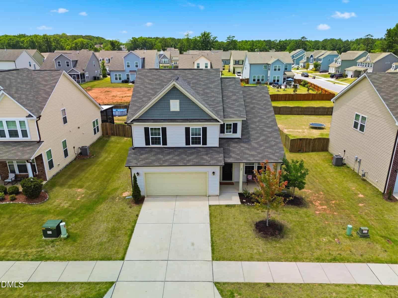 A two-story suburban house with gray siding and a covered porch, situated on a green lawn with a driveway and garage. Neighboring houses and a fenced backyard are visible under a blue sky with scattered clouds.