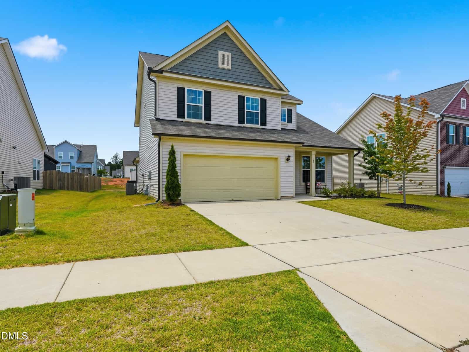 Two-story suburban house with beige siding, gray accents, and a double garage. The front yard has green grass, a small tree, and a wide driveway under a clear blue sky.