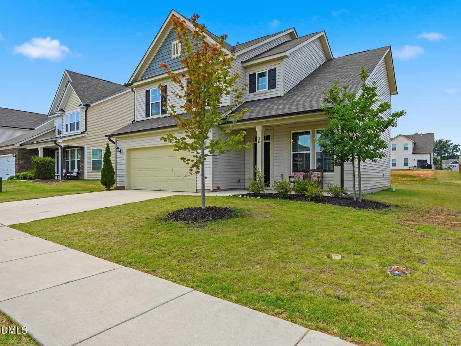 Two-story suburban house with a beige exterior, attached two-car garage, and a small front porch. The front yard has green grass, young trees, and landscaping near the entrance. A sidewalk runs along the street.