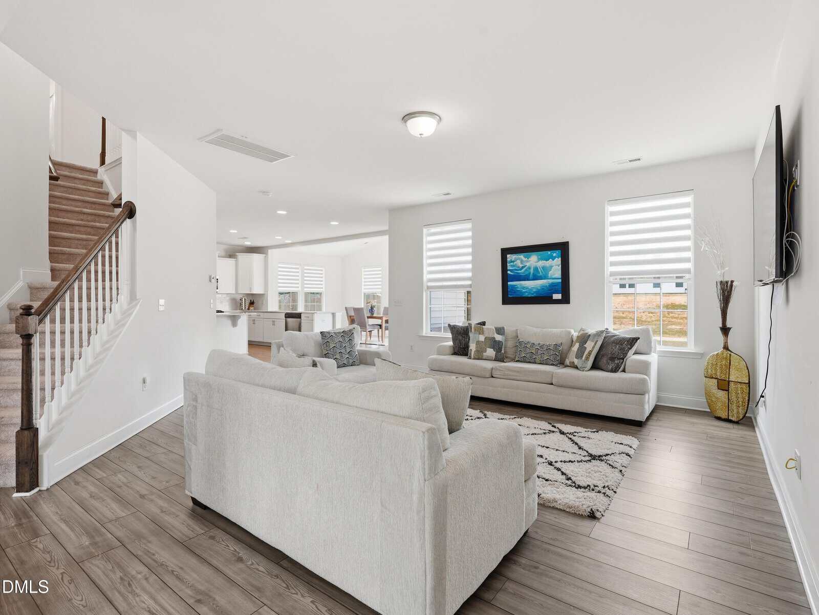 Modern living room with two light sofas, a rug, TV, framed art, and large windows. An open kitchen and dining area are visible, and a staircase leads upstairs. Wood flooring throughout. Bright, airy space.