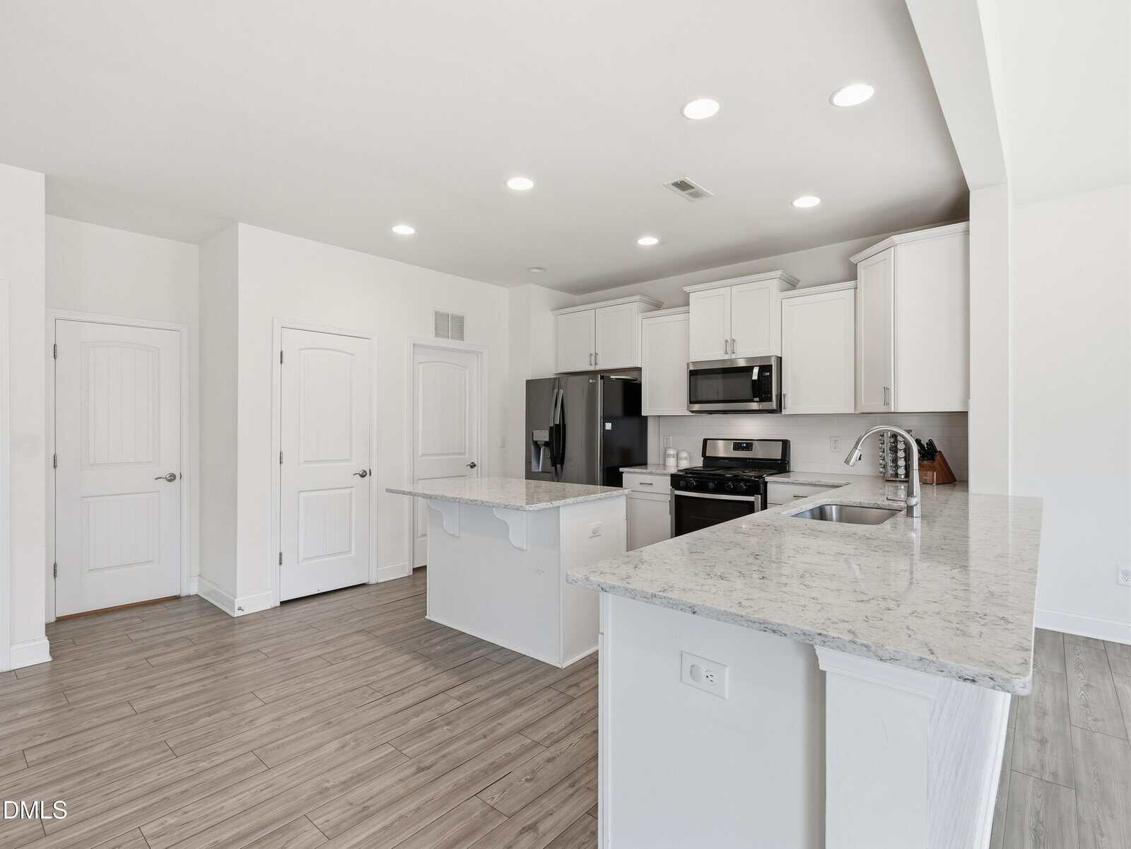 Modern kitchen with white cabinets, stainless steel appliances, marble countertops, and light wood flooring. Two islands provide extra workspace. Walls and doors are white, creating a bright, open feel.