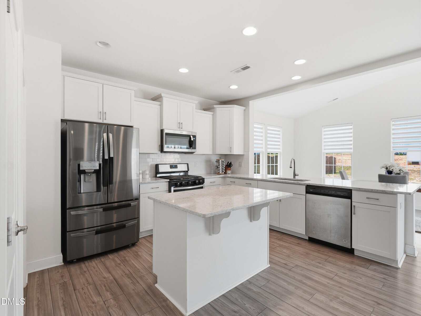 Modern white kitchen with stainless steel appliances, an island with marble countertop, white cabinets, tile backsplash, wood-look flooring, and windows allowing natural light into the space.