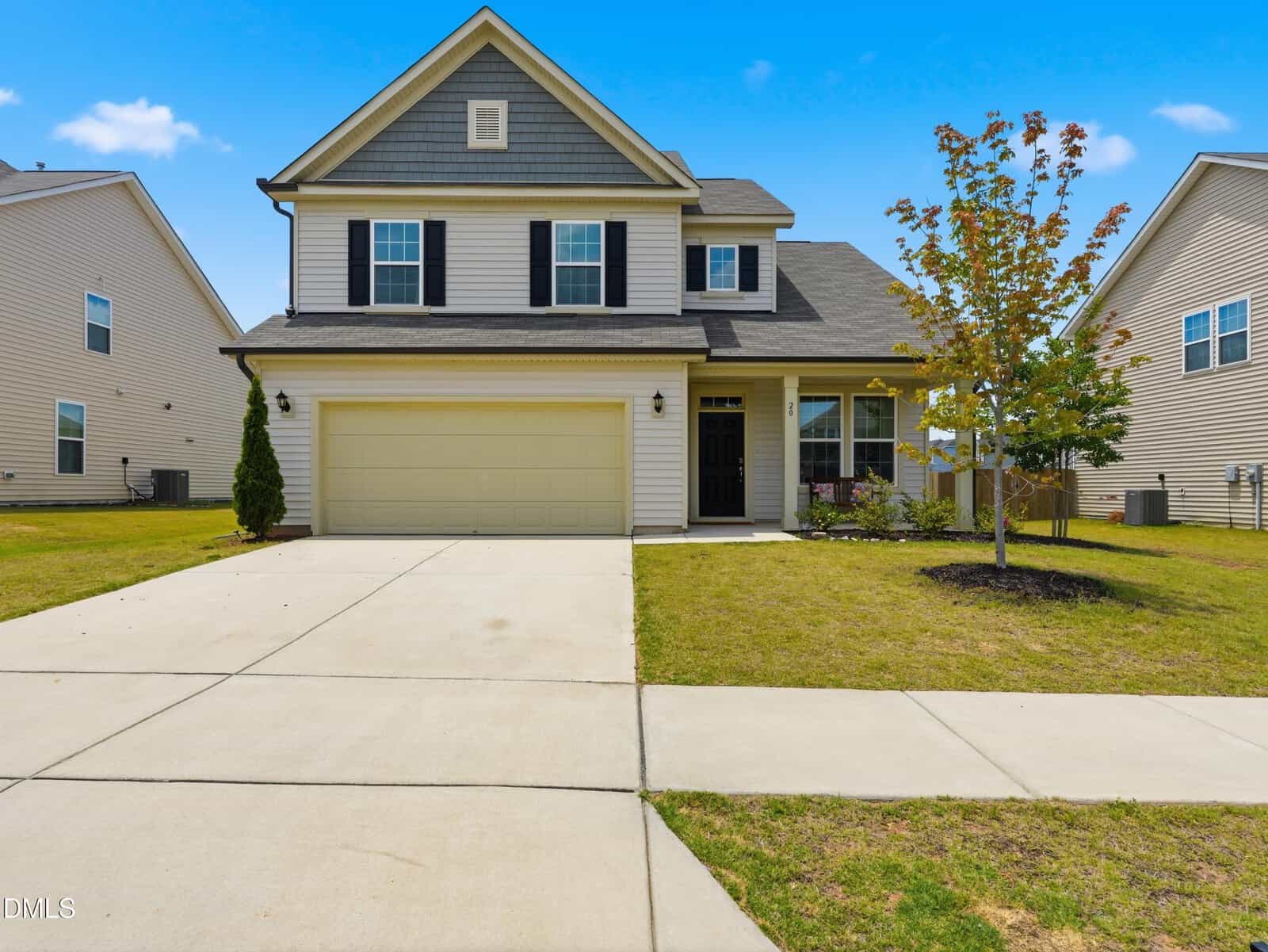 Two-story suburban house with gray siding and black shutters, attached two-car garage, concrete driveway, small front yard, single young tree, and neighboring homes on both sides under a clear blue sky.