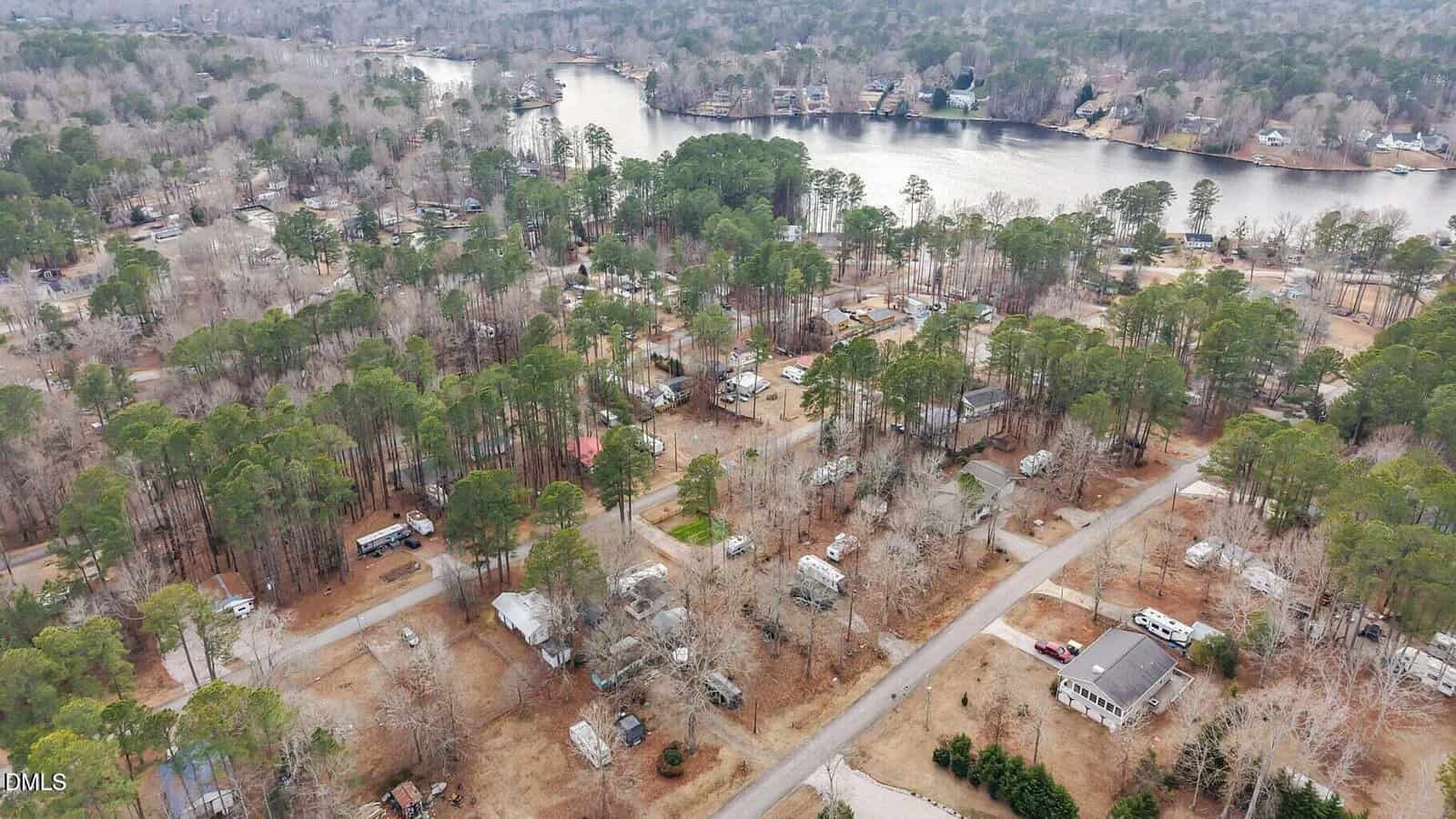 Aerial view of a wooded residential neighborhood with scattered houses and trailers, set near a lake with trees and more homes along the shoreline. Roads and driveways wind through the community.