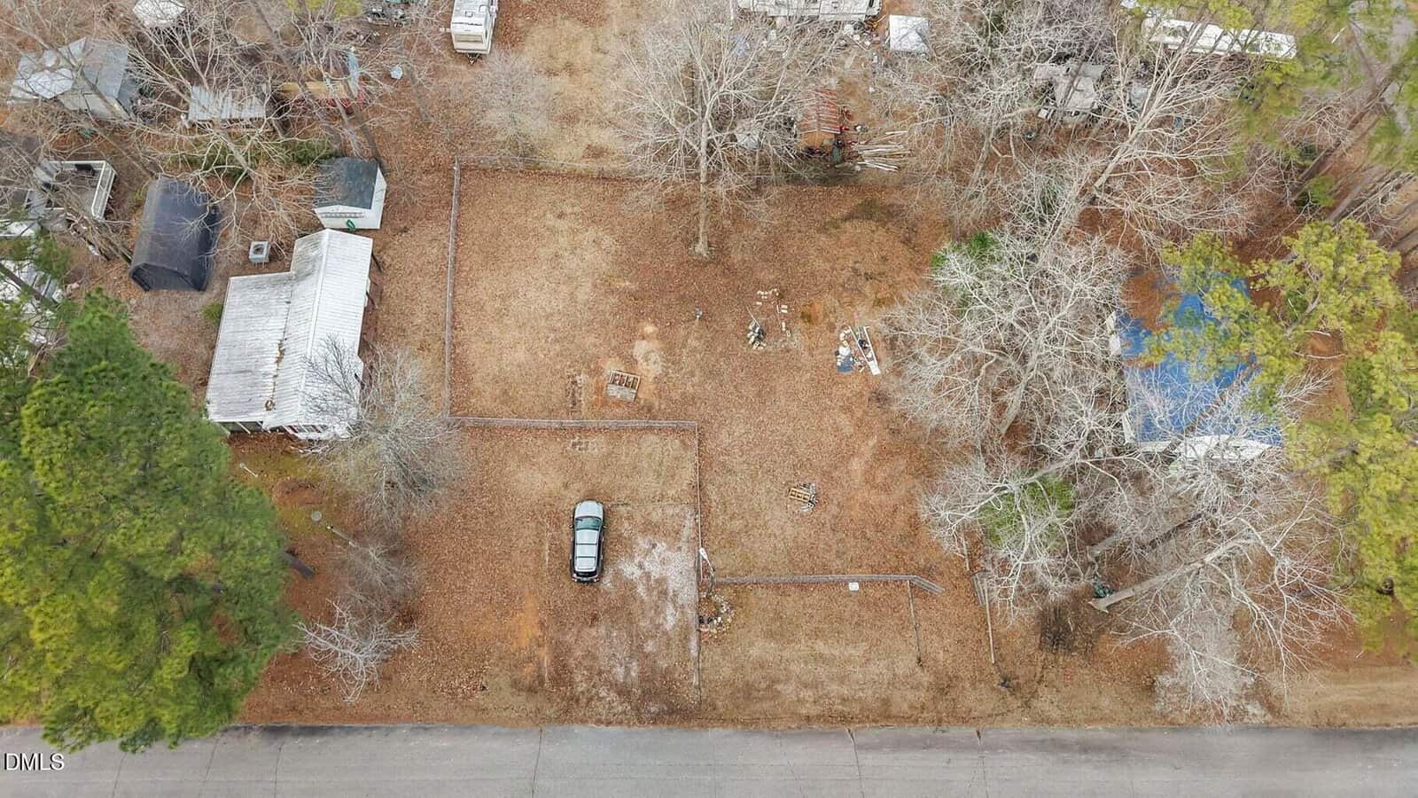 Aerial view of a rectangular, mostly bare lot with one parked car, bordered by trees and fences, and surrounded by houses and scattered outdoor items. The ground is mostly brown with sparse vegetation.