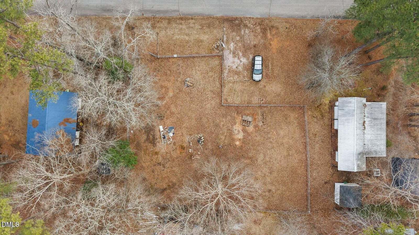 Aerial view of a rectangular, fenced yard with dry grass, scattered objects, a parked car near the top, a small blue-roofed building on the left, and a gray-roofed house with an outbuilding on the right.