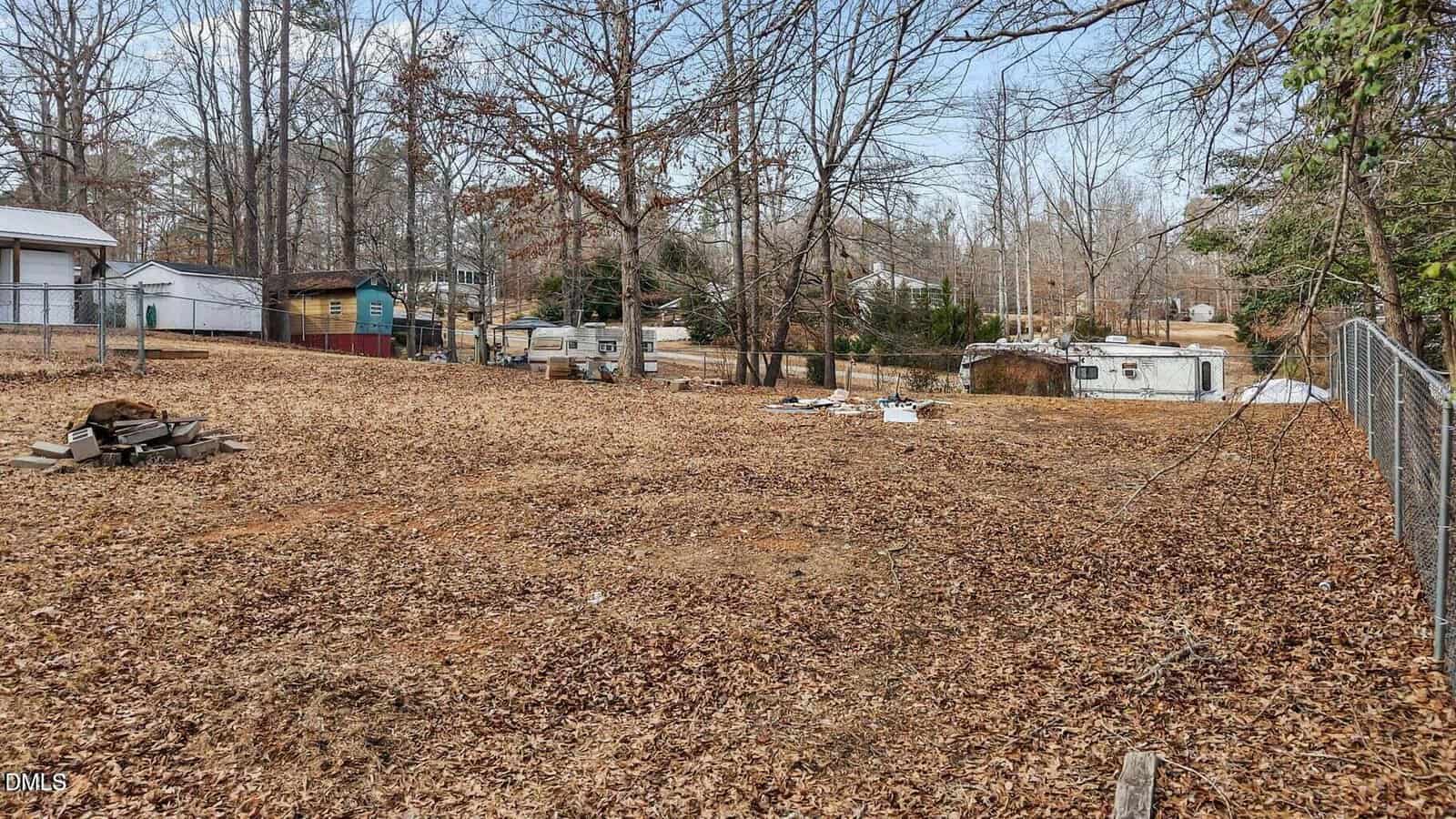 A large, leaf-covered, fenced backyard with scattered trees, sheds, and outbuildings in the background. There are some scattered items and a small fire pit area on the left side. Houses are visible in the distance.