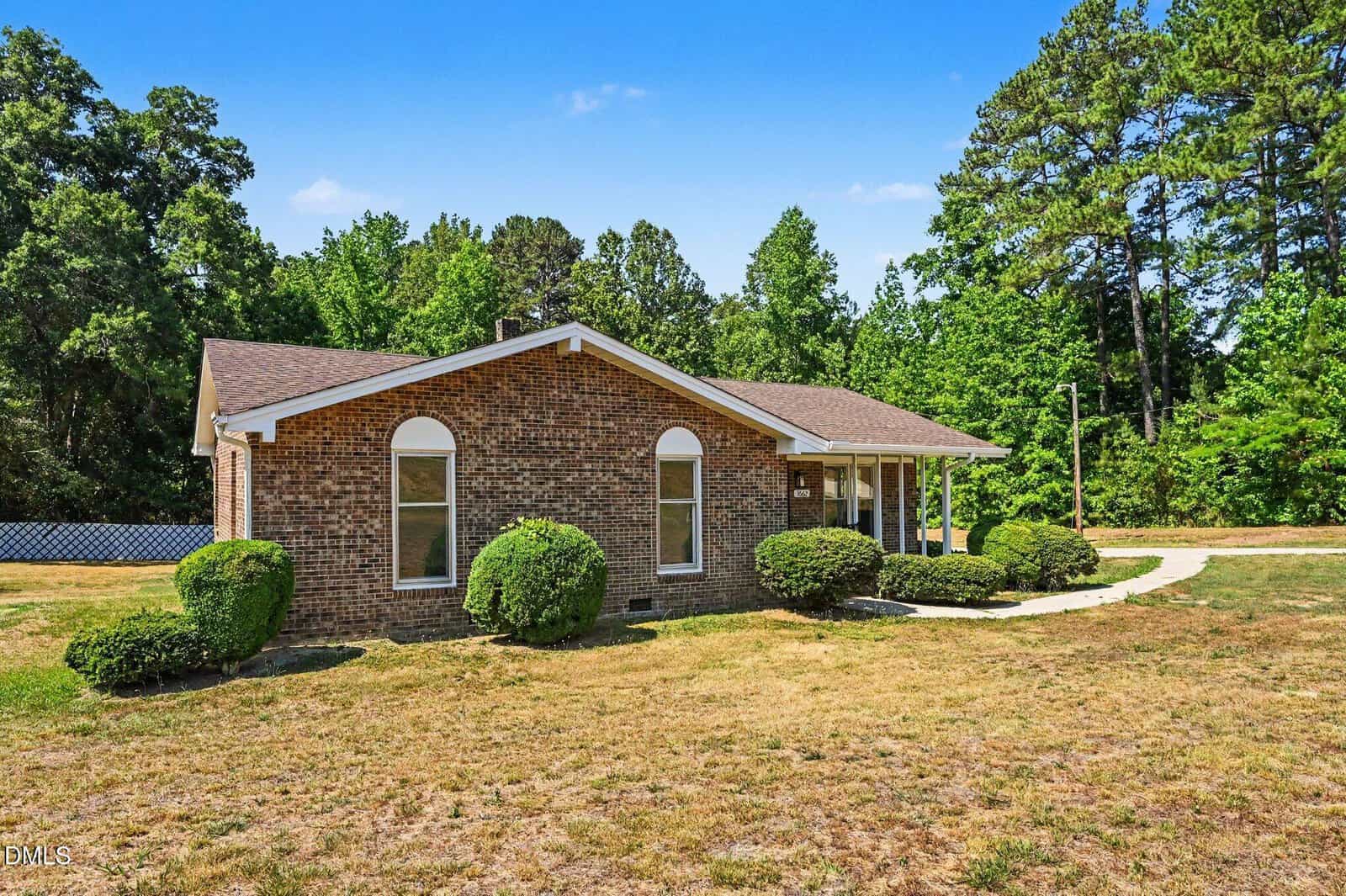A single-story brick house with arched windows, surrounded by green bushes and trees, sits on a grassy lawn under a clear blue sky.