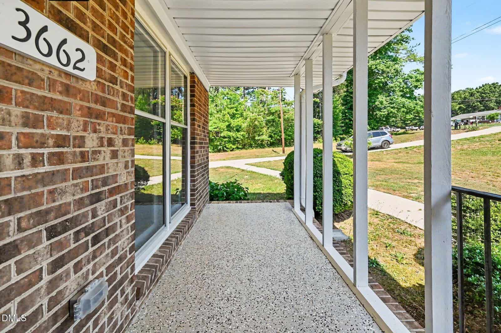 A covered front porch with white pillars, a brick wall, and the house number 3662. The porch overlooks a grassy yard with bushes, trees, and a sidewalk curving toward a quiet suburban street.