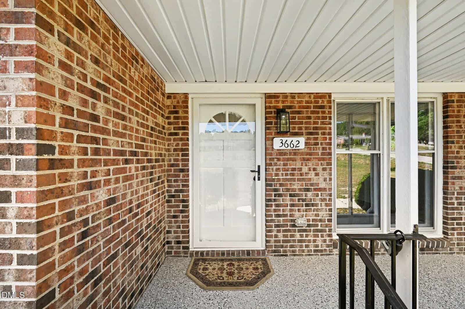 A brick house entryway with a white door, an address sign displaying "3662," a welcome mat, a black outdoor wall light, and a window with closed blinds next to the door.