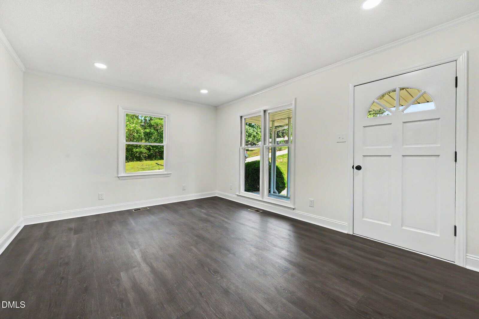 Bright, empty living room with white walls, dark wood floors, recessed lighting, two windows, and a white front door with glass panels. Outdoor greenery is visible through the windows.