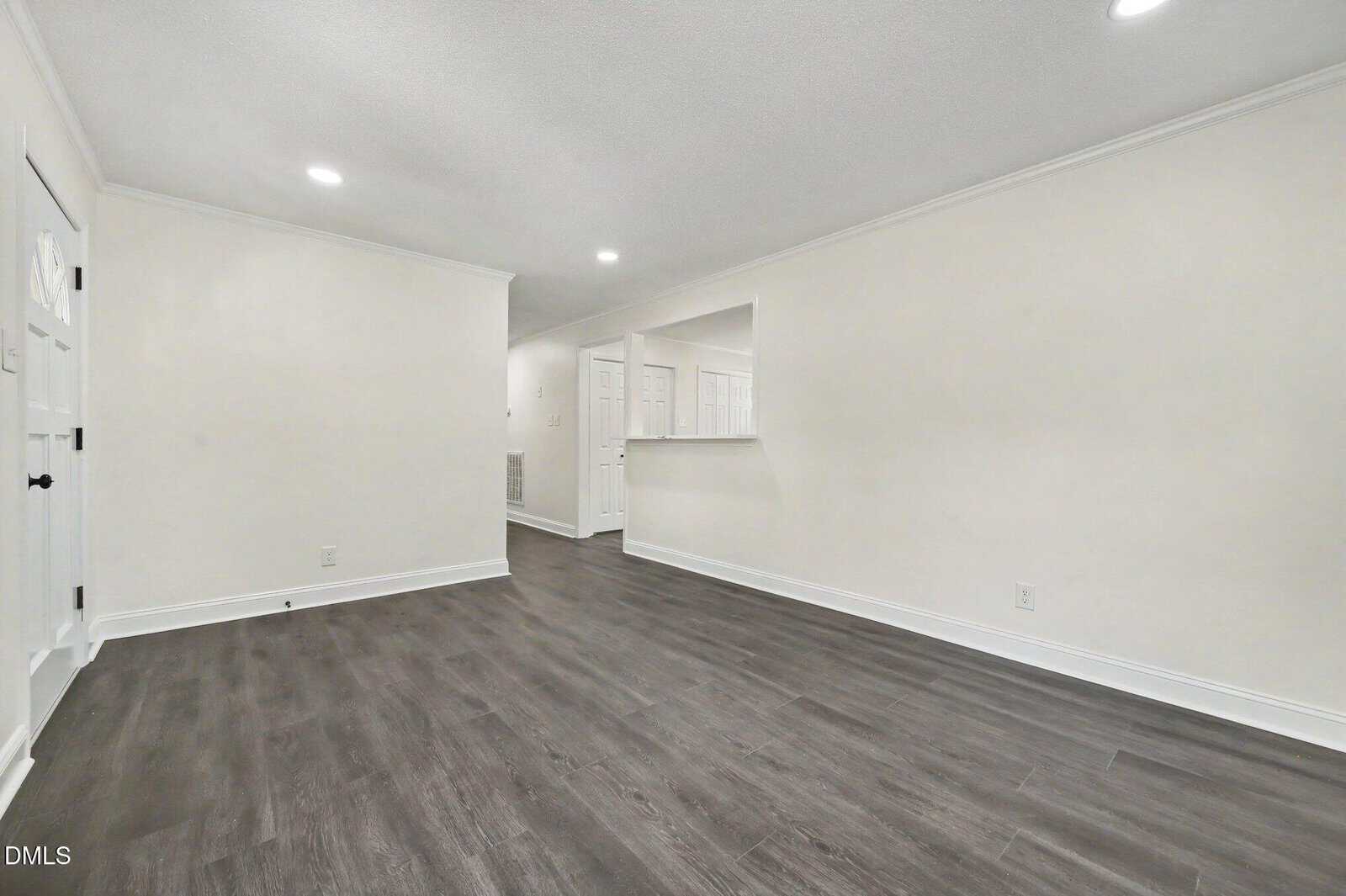 A spacious, empty room with white walls, recessed lighting, and dark wood flooring. There is a partial wall with a cutout window leading to another area and several doors in the background.