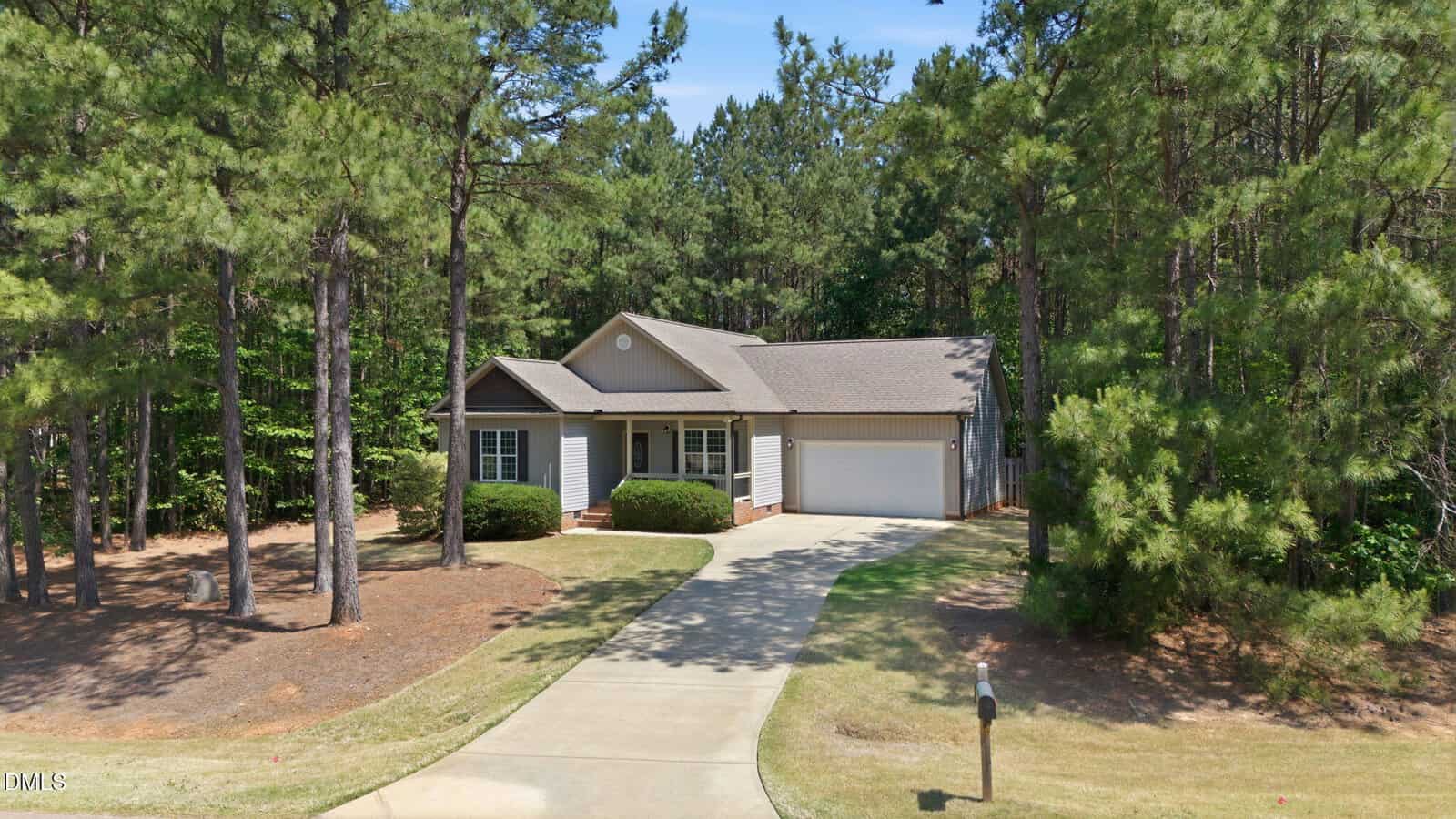 A single-story house with gray siding and a two-car garage is surrounded by tall pine trees and greenery. A curved driveway leads to the front entrance, and a mailbox stands near the street.