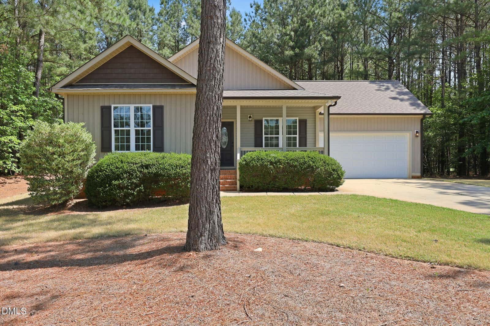 Single-story beige house with dark brown gable accents, white trim, a front porch, and attached two-car garage, surrounded by trees and shrubs, with a driveway and pine straw-covered yard in the foreground.