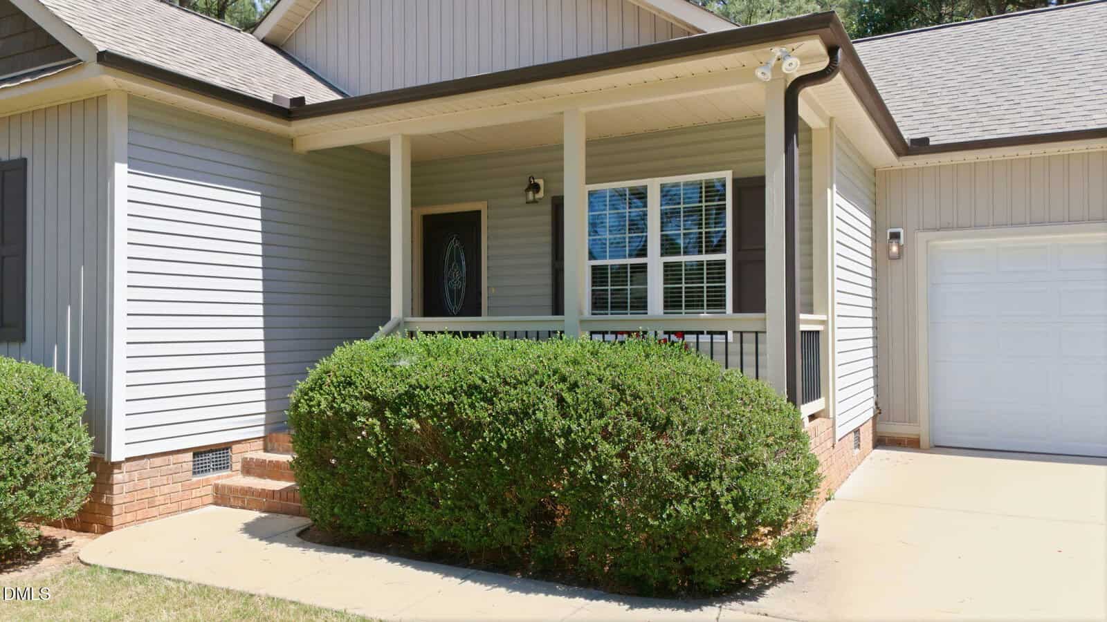 Single-story house with light gray siding, a covered front porch with black railing, brick steps, white trim, shrubs in front, and an attached garage with a white door.