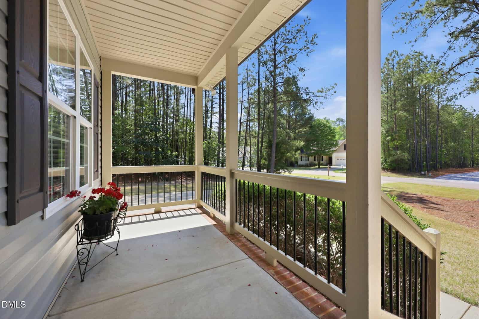 A covered front porch with black railings, potted red flowers, and a view of trees and a quiet suburban street under a clear blue sky.