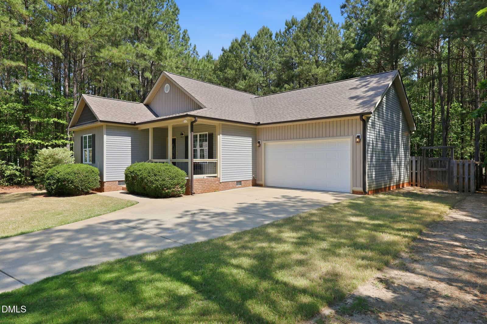 Single-story gray house with a front porch, white trim, and attached two-car garage, surrounded by trees and a well-maintained yard with a paved driveway.