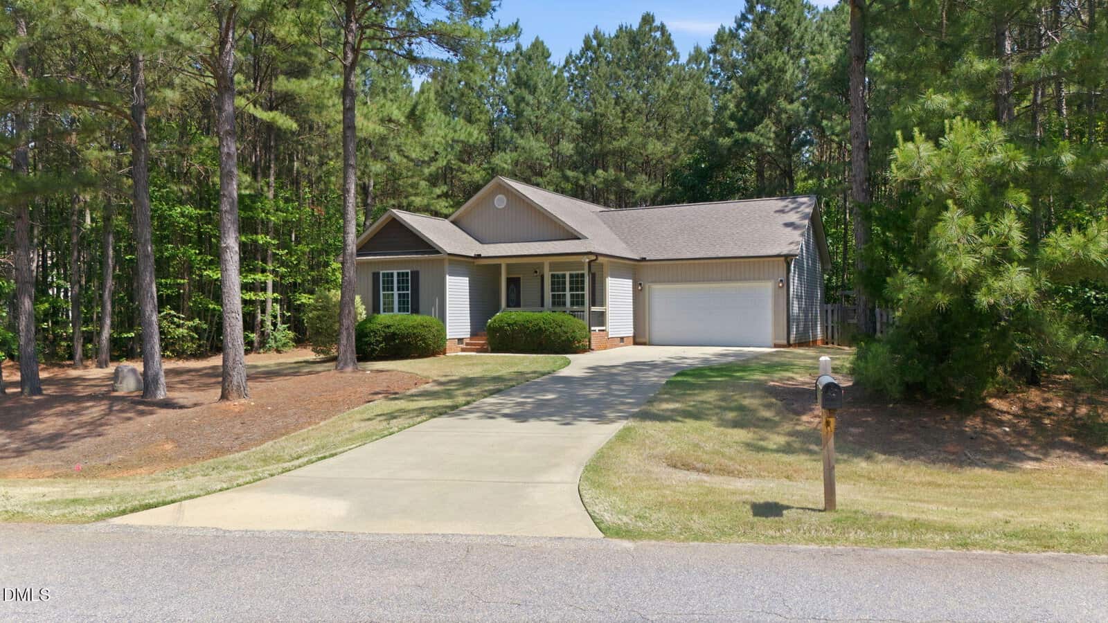 Single-story house with gray siding, white trim, and an attached garage. Surrounded by tall pine trees, it has a long driveway leading to the garage and a green lawn in front.