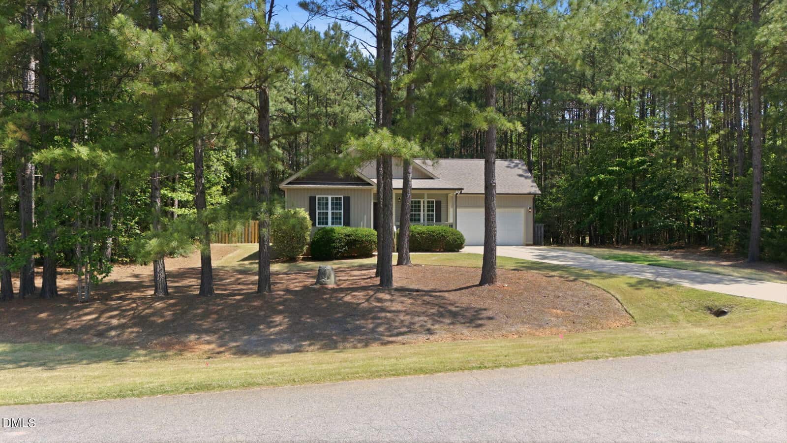 A small, single-story house with a front porch and garage is surrounded by tall pine trees and bushes, with a driveway and lawn in a wooded neighborhood.