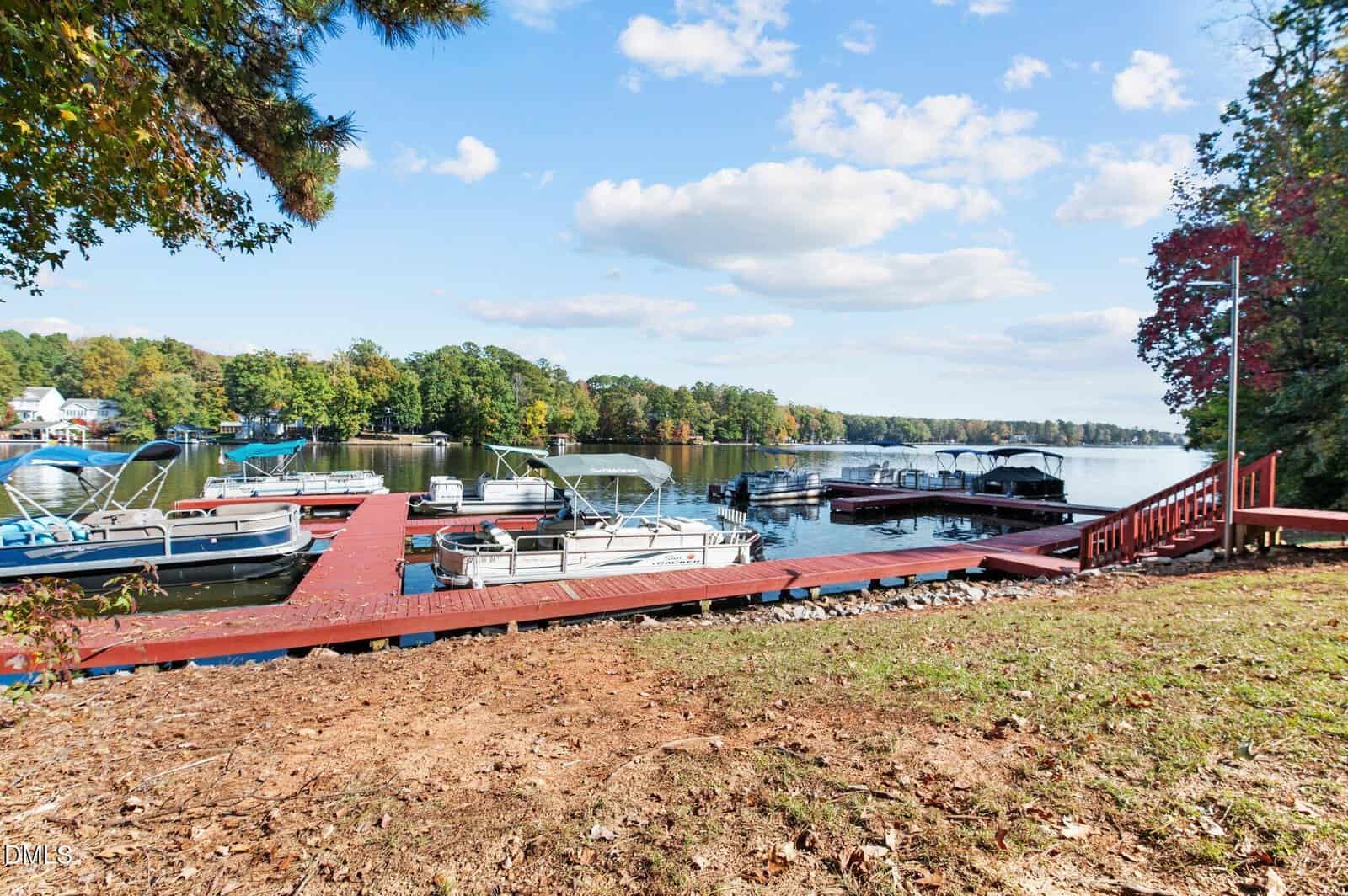Red wooden dock with several boats moored on a calm lake, surrounded by trees with green and autumn foliage, under a blue sky with scattered clouds. Steps lead from the grassy shore to the dock.