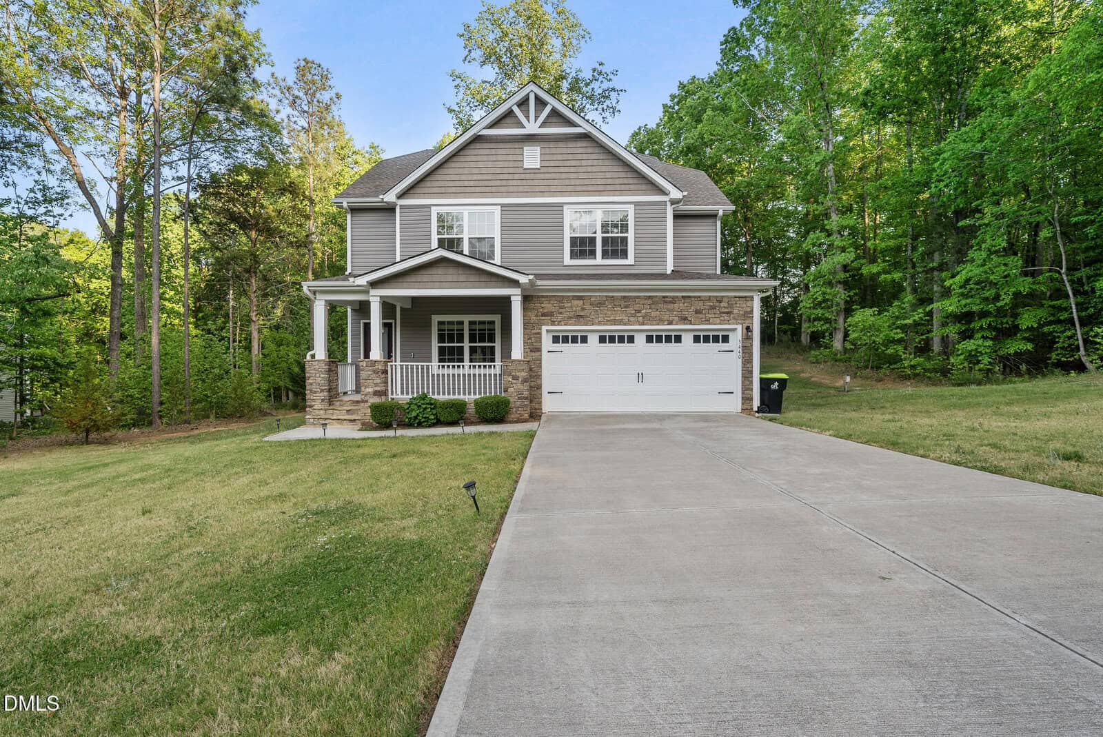 Nestled among green trees at 3440 Lilac Lane in Wake Forest, this two-story suburban house features gray siding, stone accents, a covered front porch, white trim, a double garage, and a large driveway with a well-kept lawn.