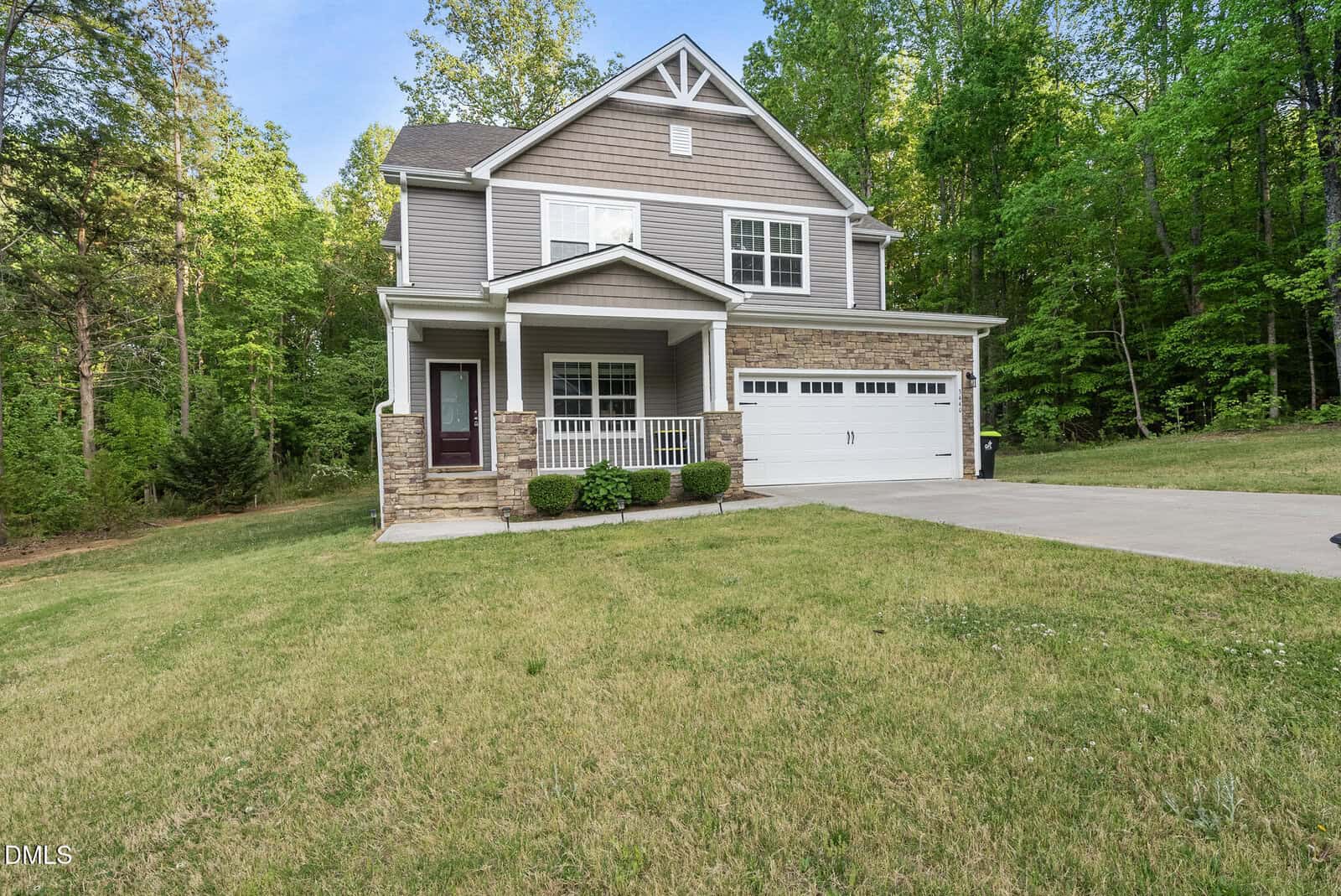 Two-story suburban house with gray siding, white trim, covered front porch, attached two-car garage, and a neatly landscaped yard, surrounded by tall green trees and a concrete driveway.