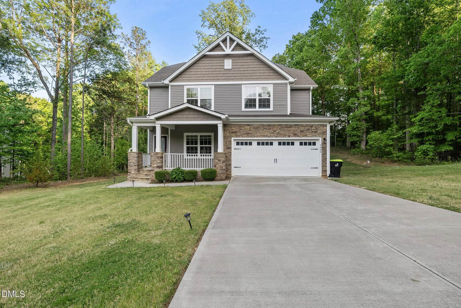 Two-story suburban house with gray siding, stone accents, white trim, and a covered front porch. A large driveway leads to a two-car garage, and the home is surrounded by a lawn and trees.