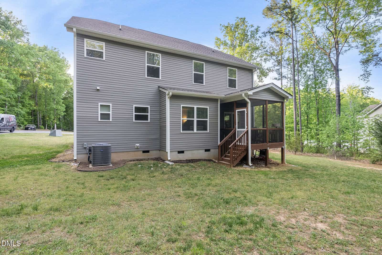 A two-story house with gray siding, several windows, and a screened-in porch with stairs leading to a grassy backyard bordered by trees.