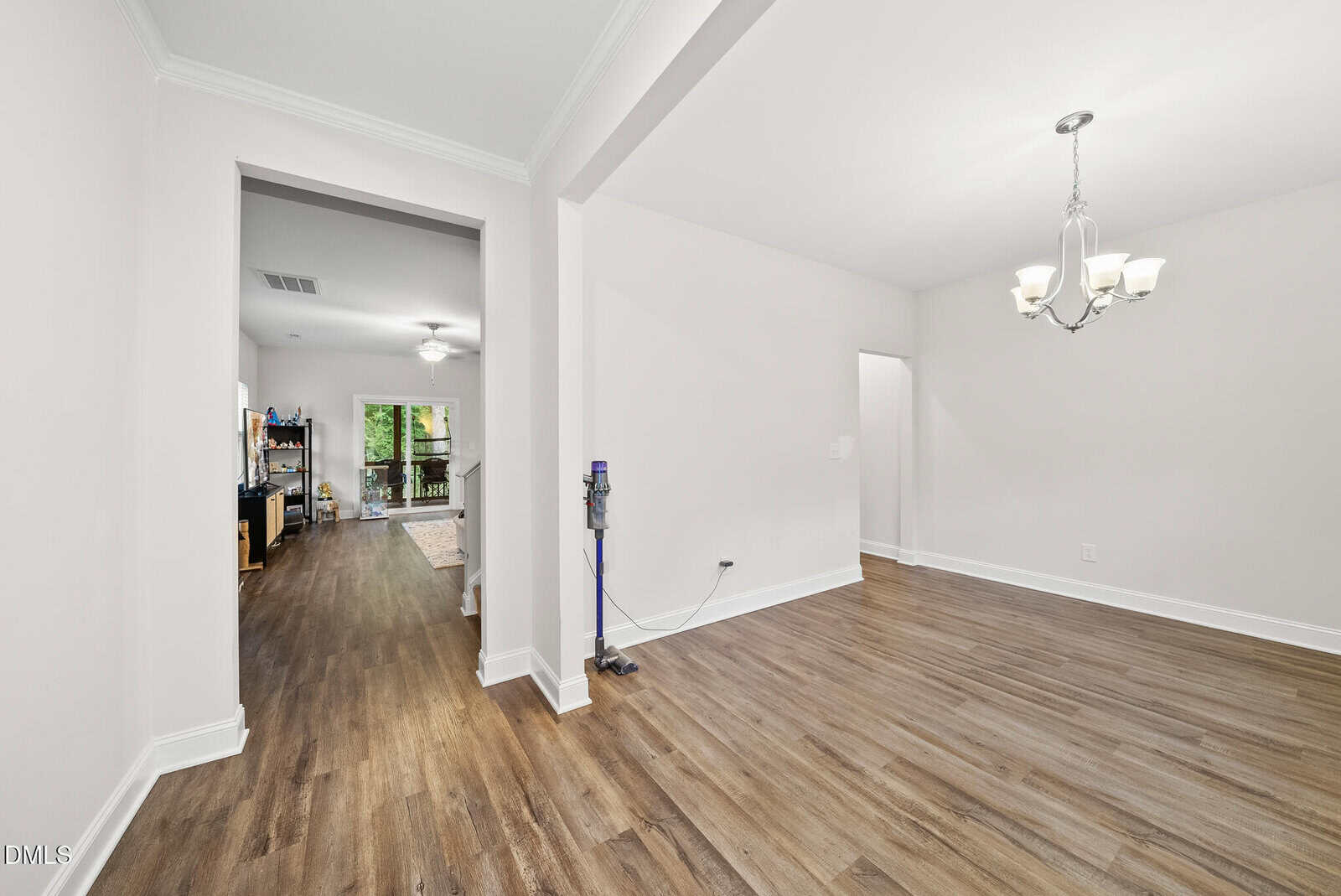 Bright, empty room with light walls, wood flooring, and a chandelier; hallway leads to a living area with shelves and a view of greenery through glass doors; a vacuum is placed against one wall.