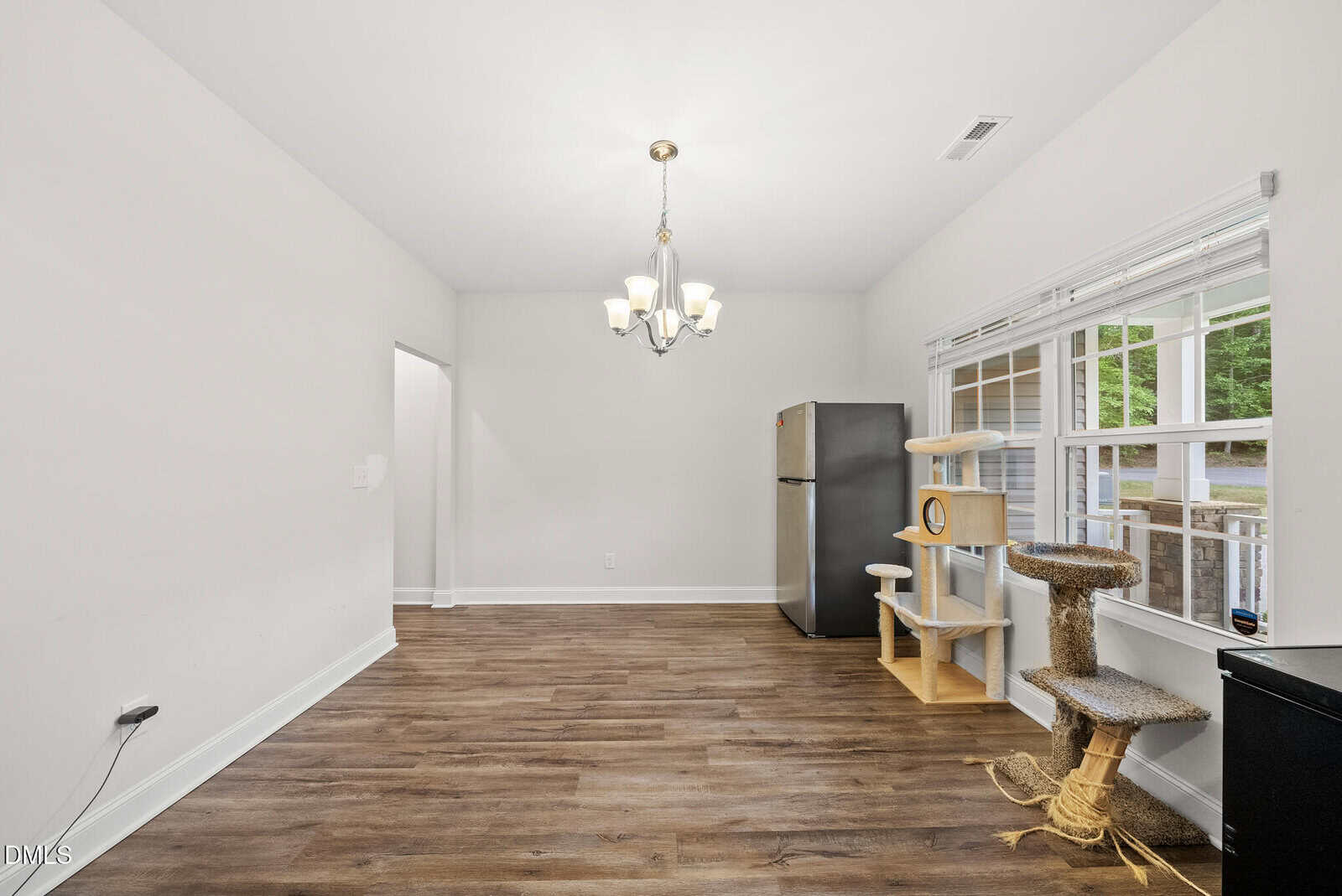 A bright room with wood floors, a silver fridge, cat tree, and large windows. A modern chandelier hangs from the ceiling, and natural light fills the space.