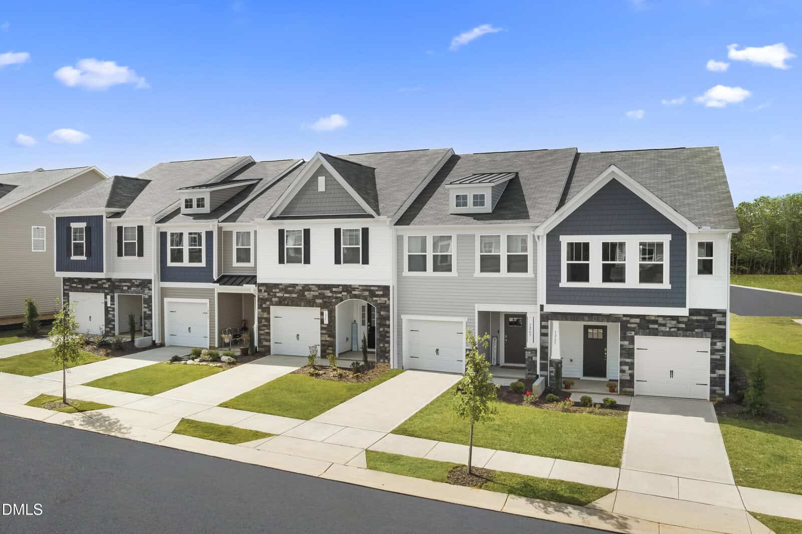 A row of modern two-story townhouses with attached garages, neatly landscaped front yards, and a clean sidewalk along a quiet suburban street under a blue sky with scattered clouds.