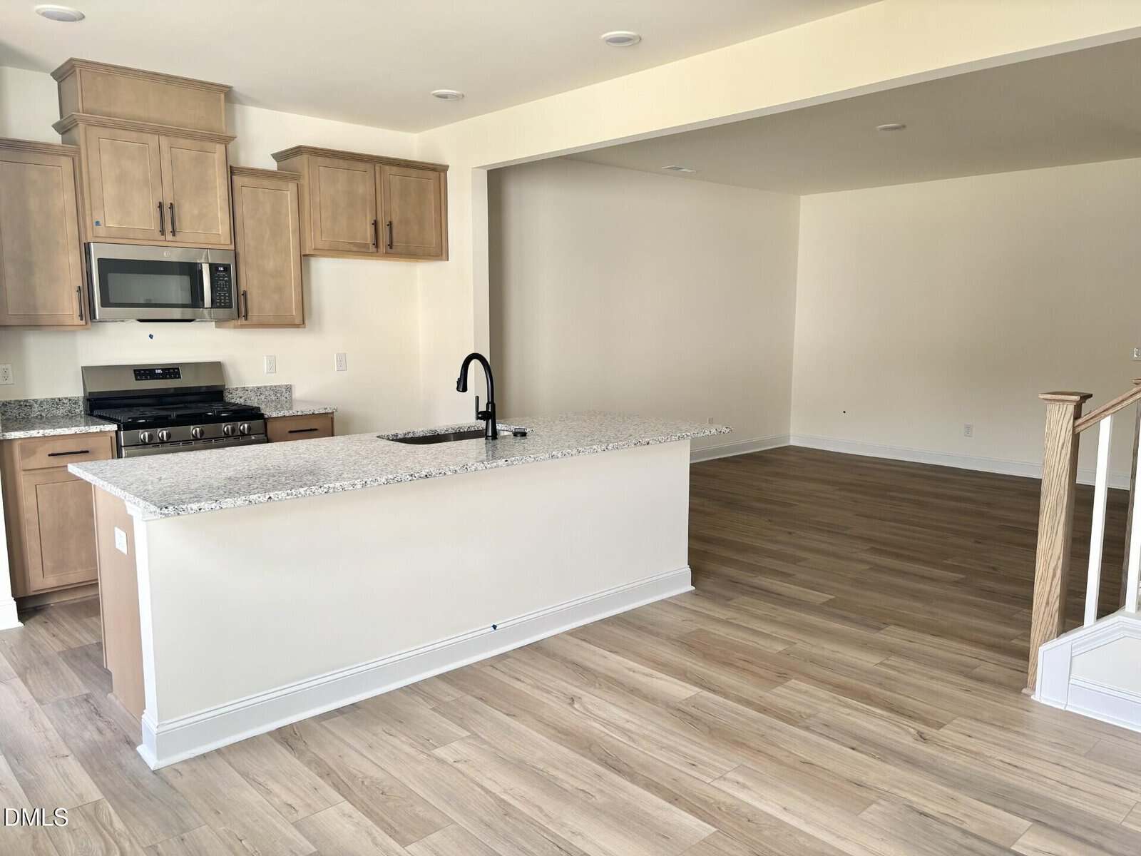 Modern kitchen with wood cabinets, granite island with a black faucet, and stainless steel appliances, opening to an empty living area with light wood floors and a partial view of stairs.