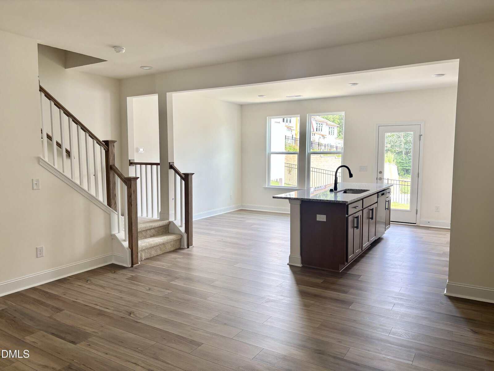 Open-concept kitchen and living area with wood floors, island with sink, large windows, white walls, and stairs leading up. Glass door opens to a sunny backyard. Natural light fills the bright, modern space.