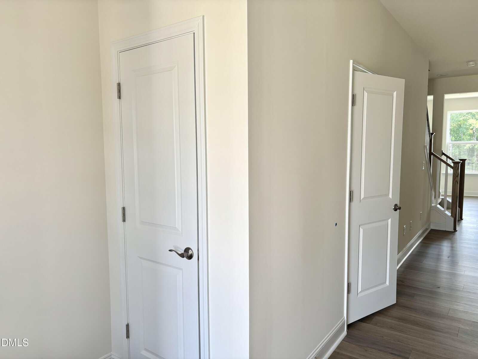 A hallway with two white doors, one closed and one open. The hallway has light-colored walls and wood flooring. Sunlight is visible through a window in the background near a staircase.
