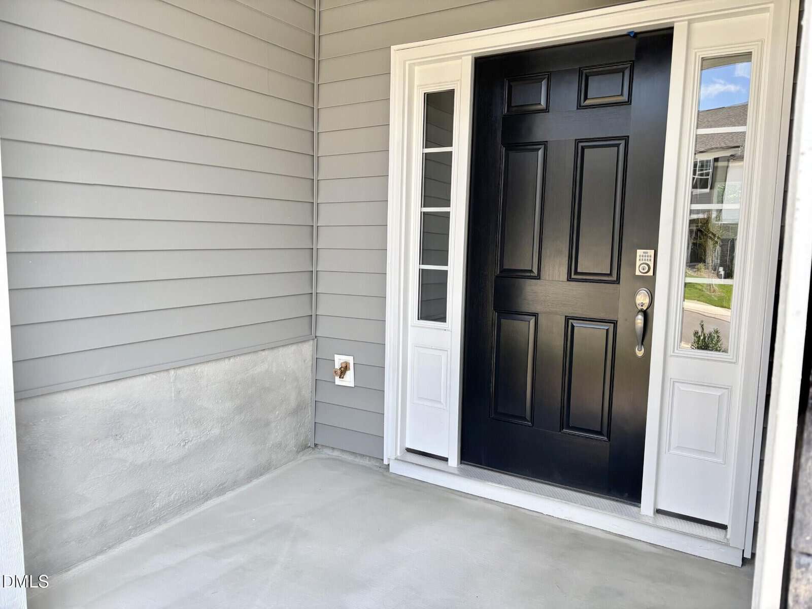 A covered front entryway with gray siding, a black front door with glass sidelights, a concrete floor, and a small wall fixture. A house and landscaping are visible through the sidelights.