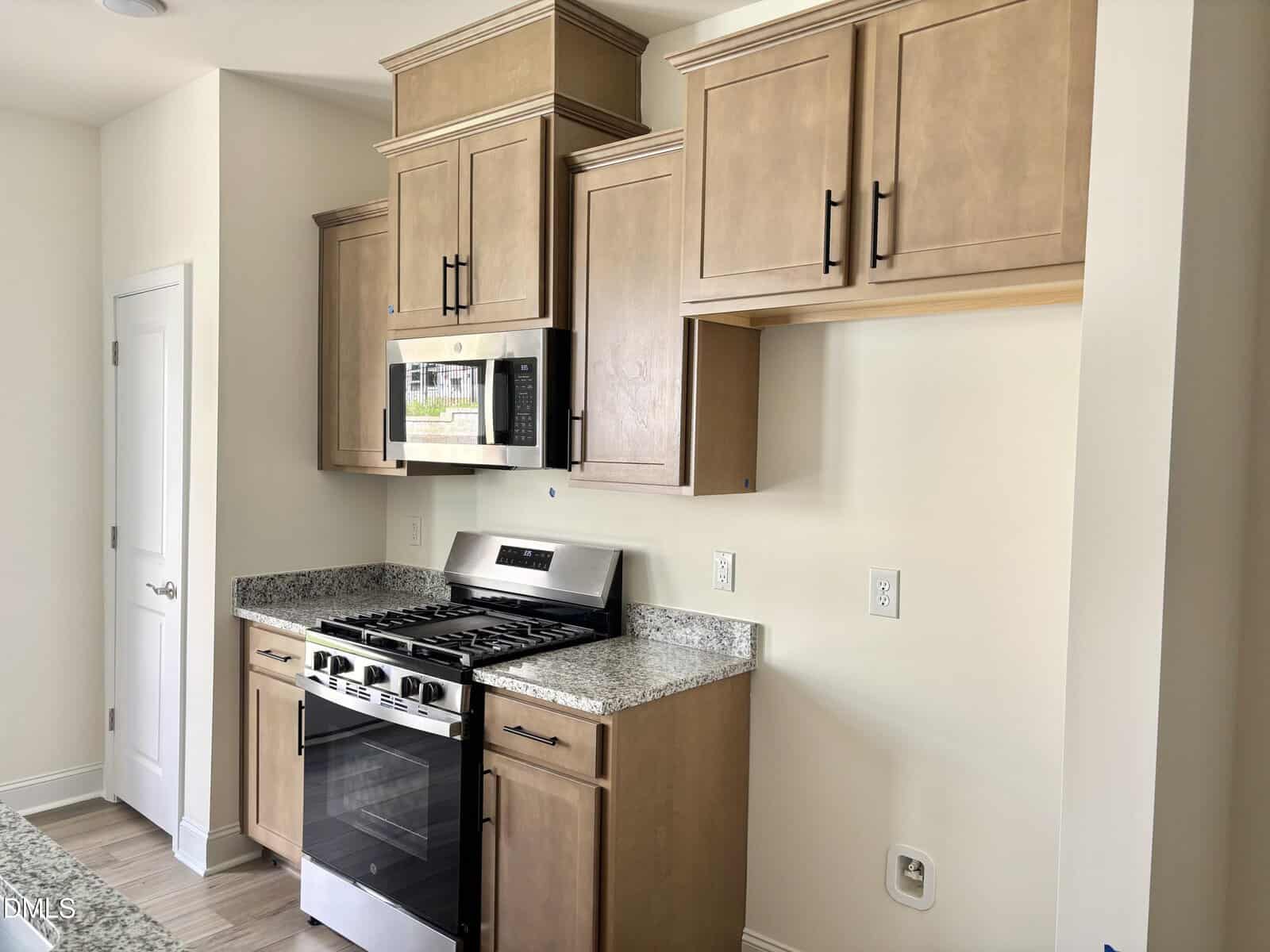 A modern kitchen with light wood cabinets, granite countertops, a stainless steel gas stove with oven, and a built-in microwave. There are electrical outlets on the wall and no items on the counters.