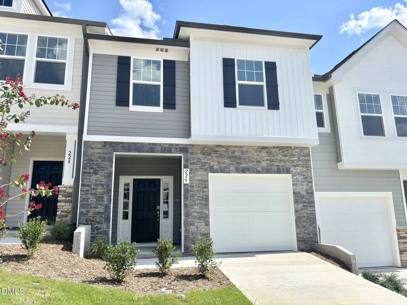 Charming two-story townhouse at 226 Alberta Drive in Franklinton, featuring gray siding, white trim, and elegant stone accents. This home offers a single-car garage, a black front door, two upper windows with stylish black shutters, and a small landscaped front yard under a clear blue sky.