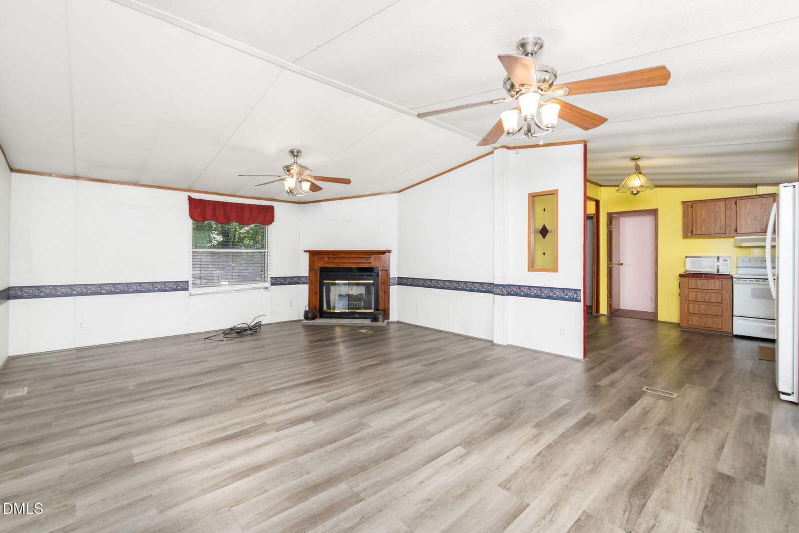 Spacious living room with light wood flooring, white walls, two ceiling fans, a fireplace, large window with a red valance, and an open view into a kitchen with yellow walls and wooden cabinets.