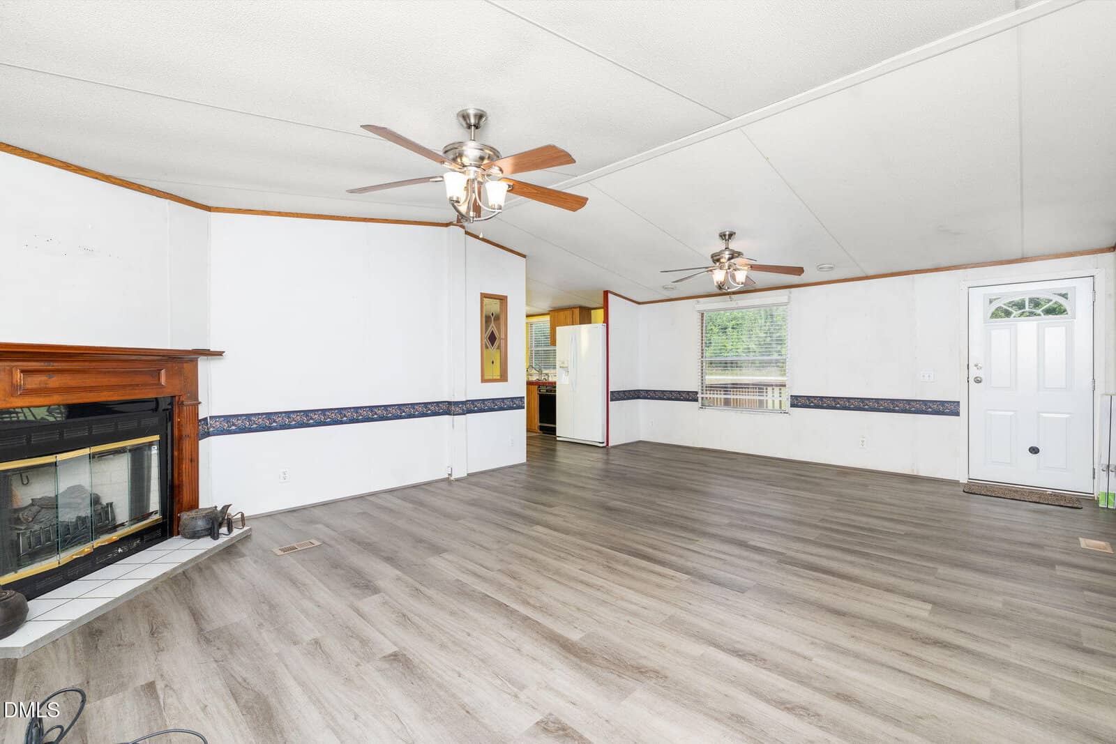 Spacious living room with light wood flooring, white walls, two ceiling fans, a fireplace with a wooden mantel, and large windows letting in natural light. A door and partial view of the kitchen are visible.