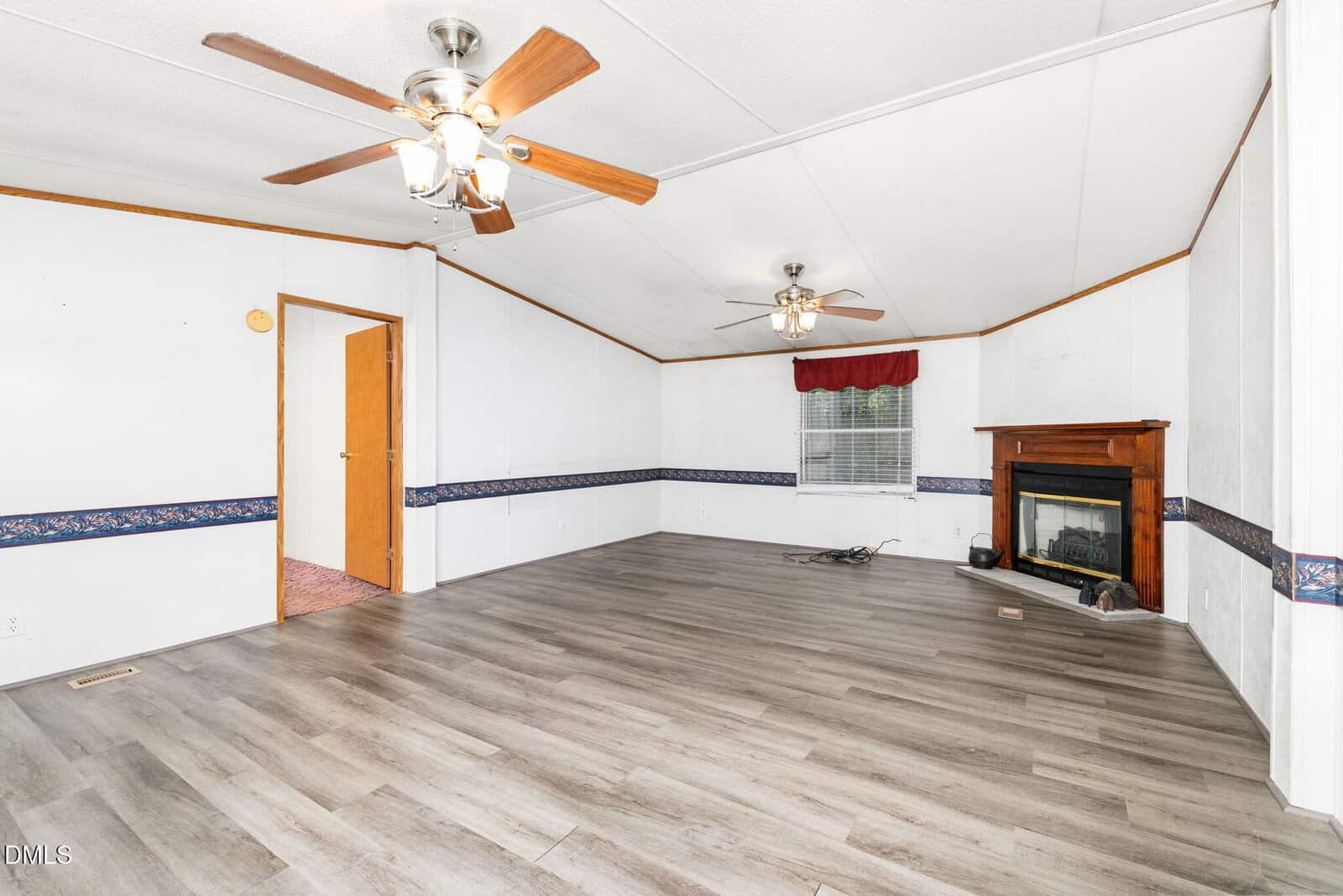Empty living room with light wood flooring, white walls, two ceiling fans, a window with a red valance, a wooden fireplace, and a doorway leading to another room. Blue patterned trim runs along the walls.