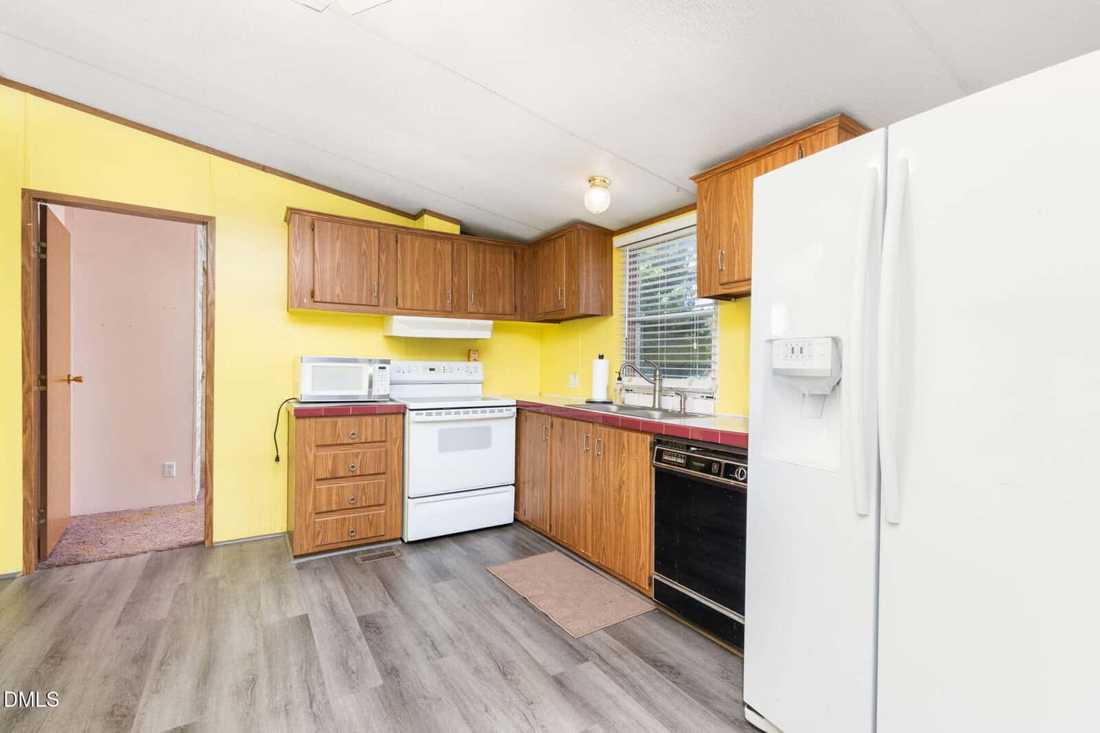 A kitchen with yellow walls, wood cabinets, white appliances, a dishwasher, and a large white refrigerator. There’s a window above the sink and gray wood-style flooring. An open doorway reveals a carpeted room.