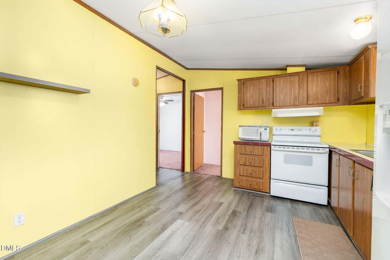 A small kitchen with yellow walls, wood cabinets, a white stove, microwave, and a fridge. Open doorways lead to other rooms; the floor is gray wood laminate. A shelf is mounted on the left wall.