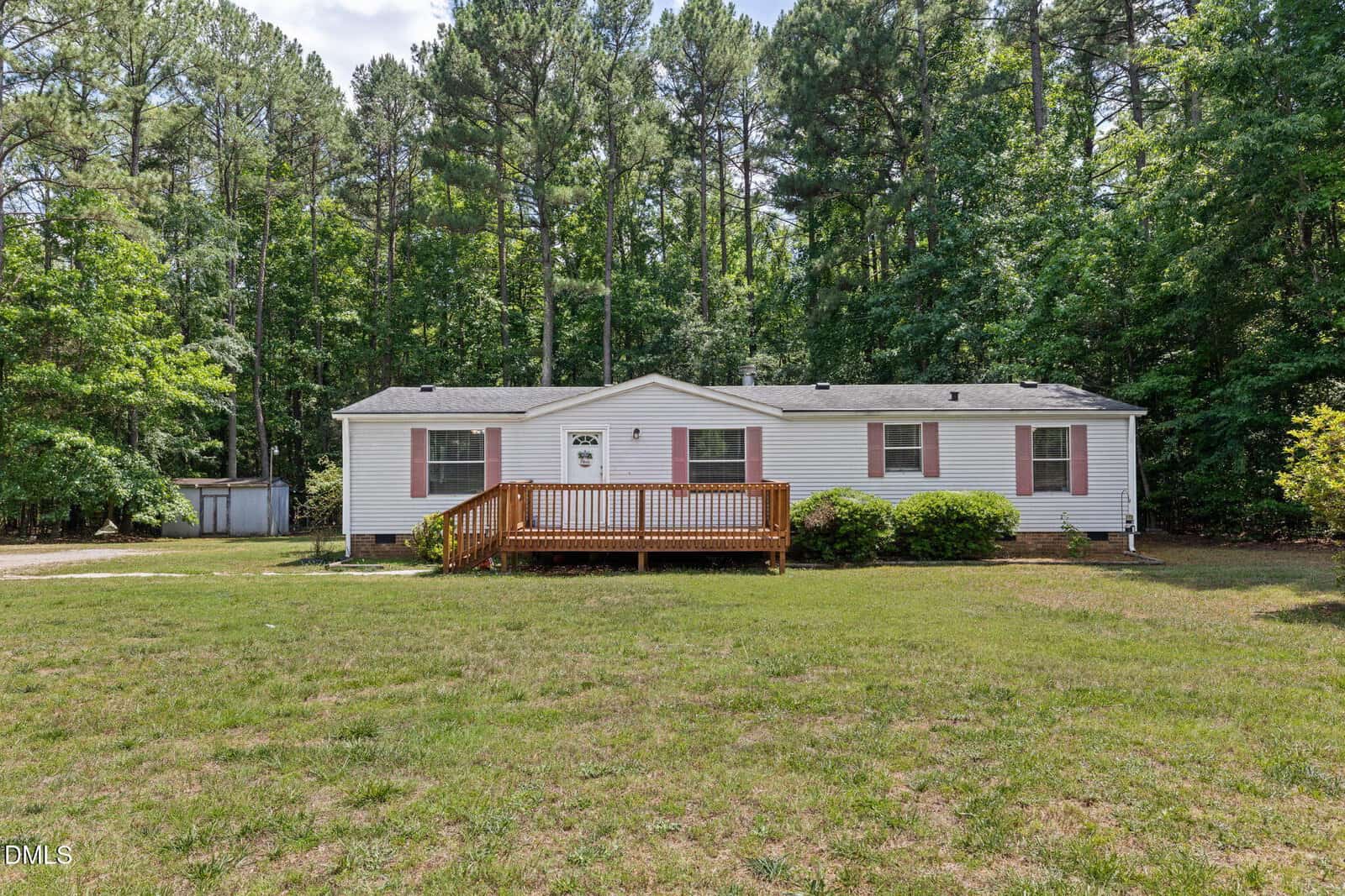 A single-story white house with a wooden front porch sits in a large grassy yard at 1232 Plum Nutty Road, Henderson, surrounded by tall pine trees and dense greenery under a partly cloudy sky.