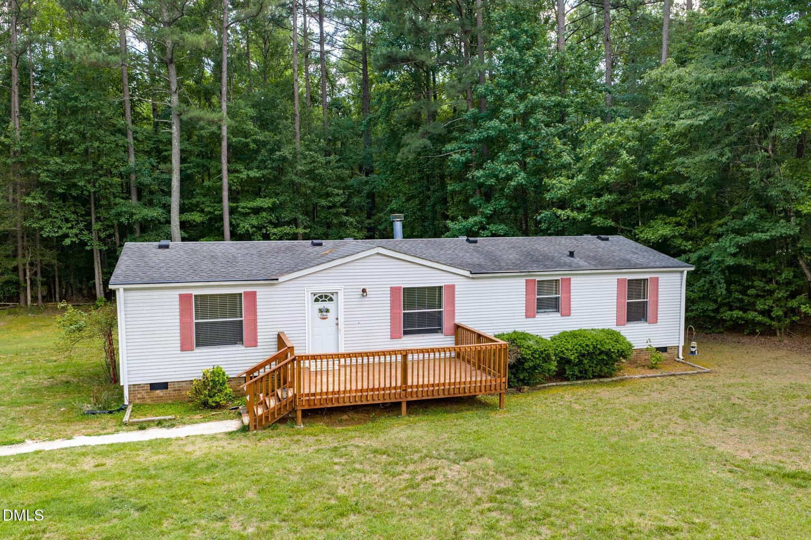 A single-story white house with black roof and pink shutters, featuring a wooden porch and steps. The house is surrounded by green grass and dense trees in the background.
